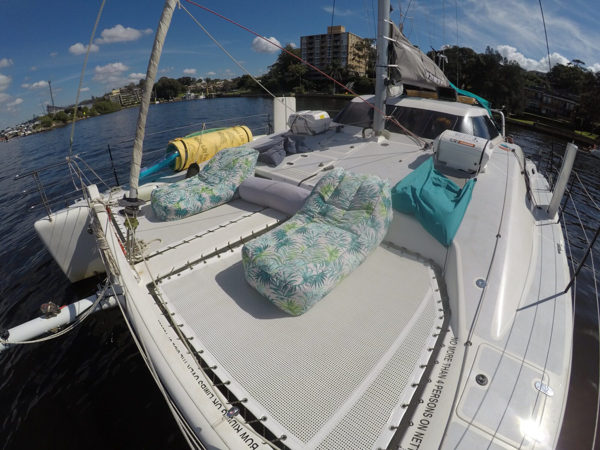 A sailboat, part of The Yacht Social Club, is docked on a calm river, featuring a spacious deck with patterned lounge chairs, teal and white cushions, and various boating equipment. The background shows a cityscape with buildings, green trees, and a clear blue sky with scattered clouds.