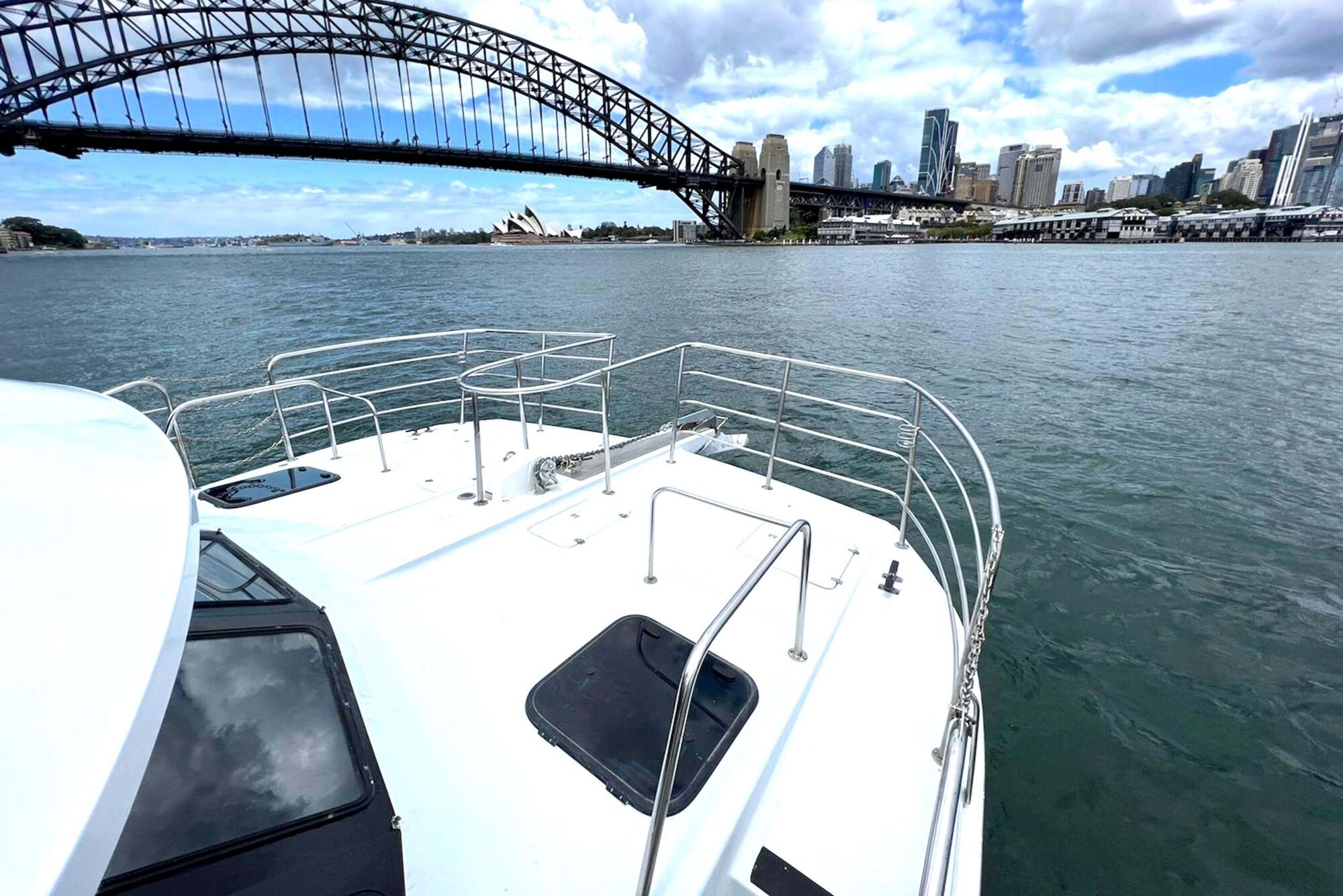 View of Sydney Harbour from the deck of a luxury yacht rental, showcasing the Sydney Harbour Bridge and the Sydney Opera House in the background, along with the city skyline. The sky is partly cloudy, and the water appears calm.