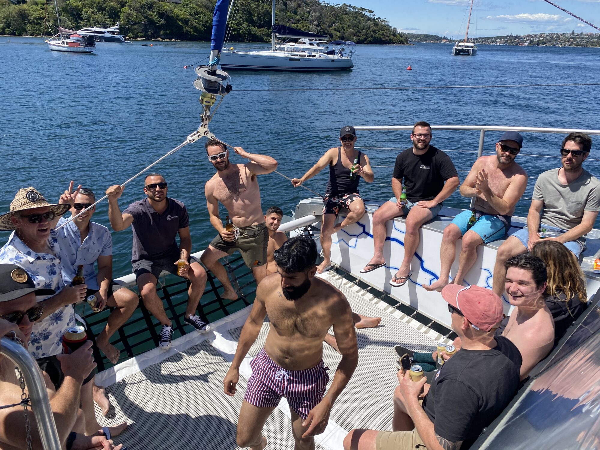 A group of people enjoying a sunny day on a boat. They are smiling, laughing, and holding drinks. Some are sitting while others stand or dance. In the background, other boats and a scenic landscape with trees and blue water are visible.