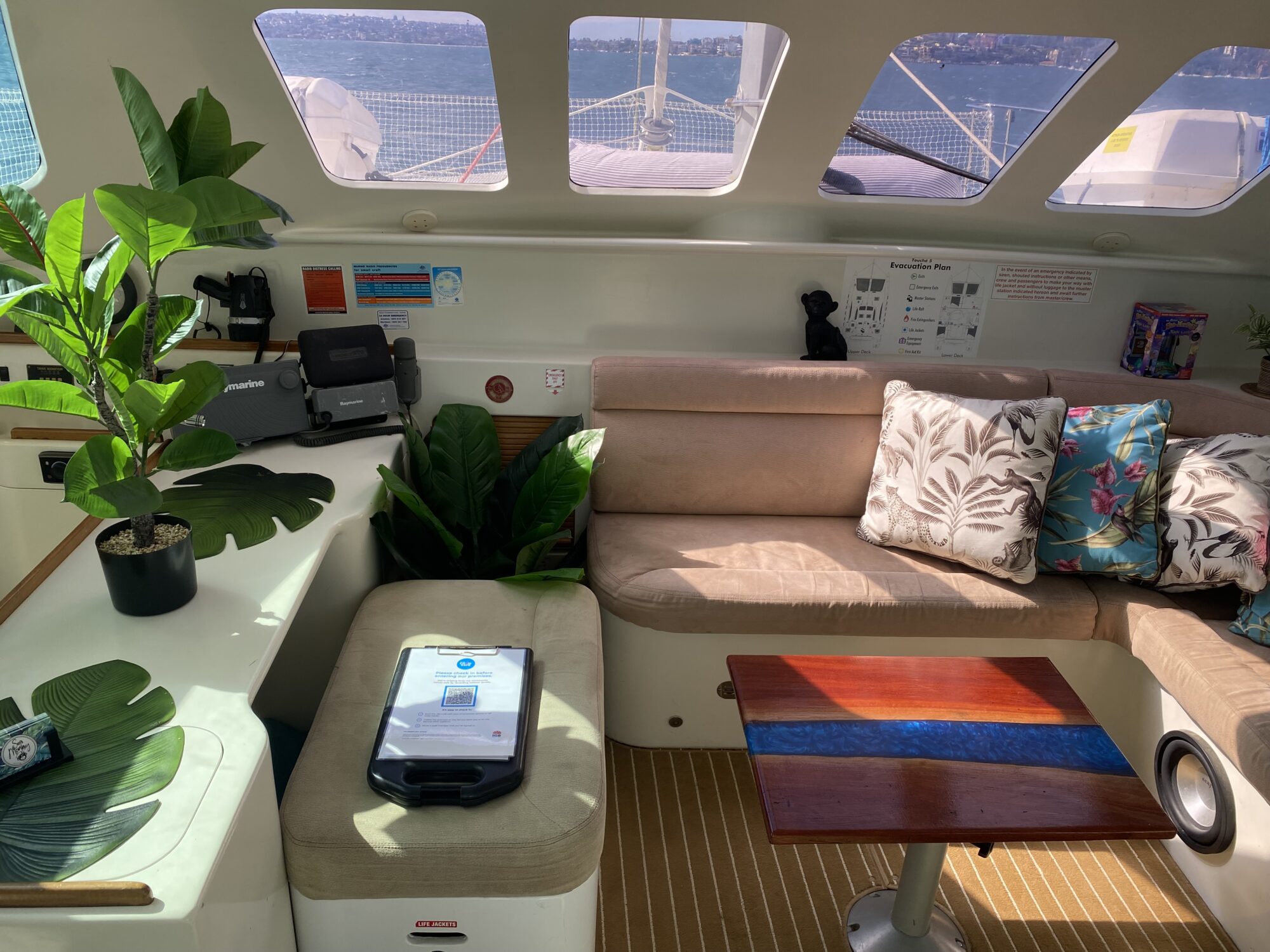 Interior of a luxury yacht's cabin with large windows offering an ocean view. The space features cushioned seating with floral pillows, a wooden table, potted plants, and navigation equipment on a counter. Sunlight streams in, creating a bright and cozy atmosphere—perfect for Boat Parties by The Yacht Social Club.