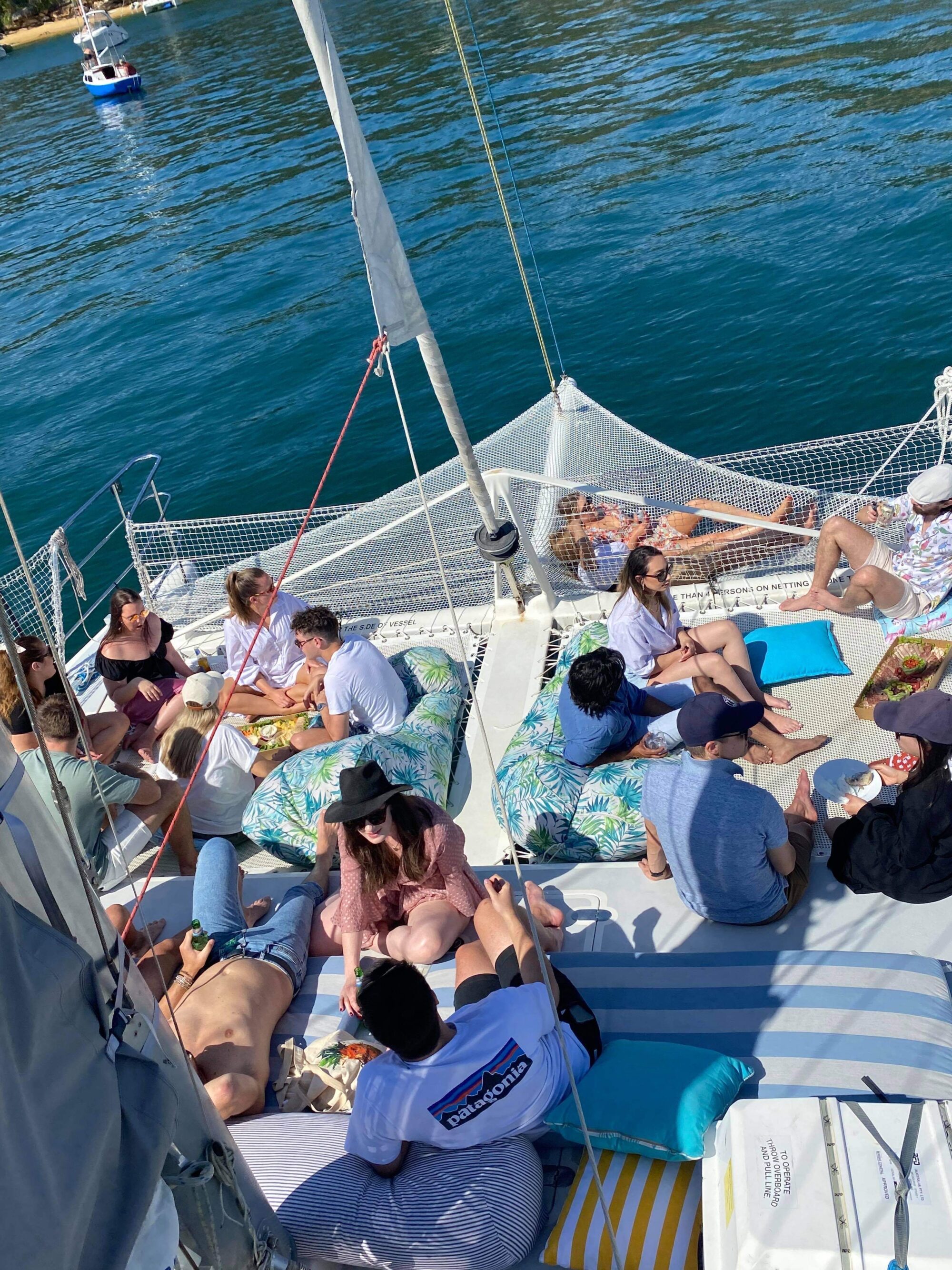 A group of people relaxing on a boat under sunny weather. Some are sitting, others are lying on cushions and pillows. The boat is anchored in a calm, blue body of water. Snacks and drinks are visible, creating a leisurely and enjoyable atmosphere reminiscent of Boat Parties by The Yacht Social Club in Sydney Harbour.