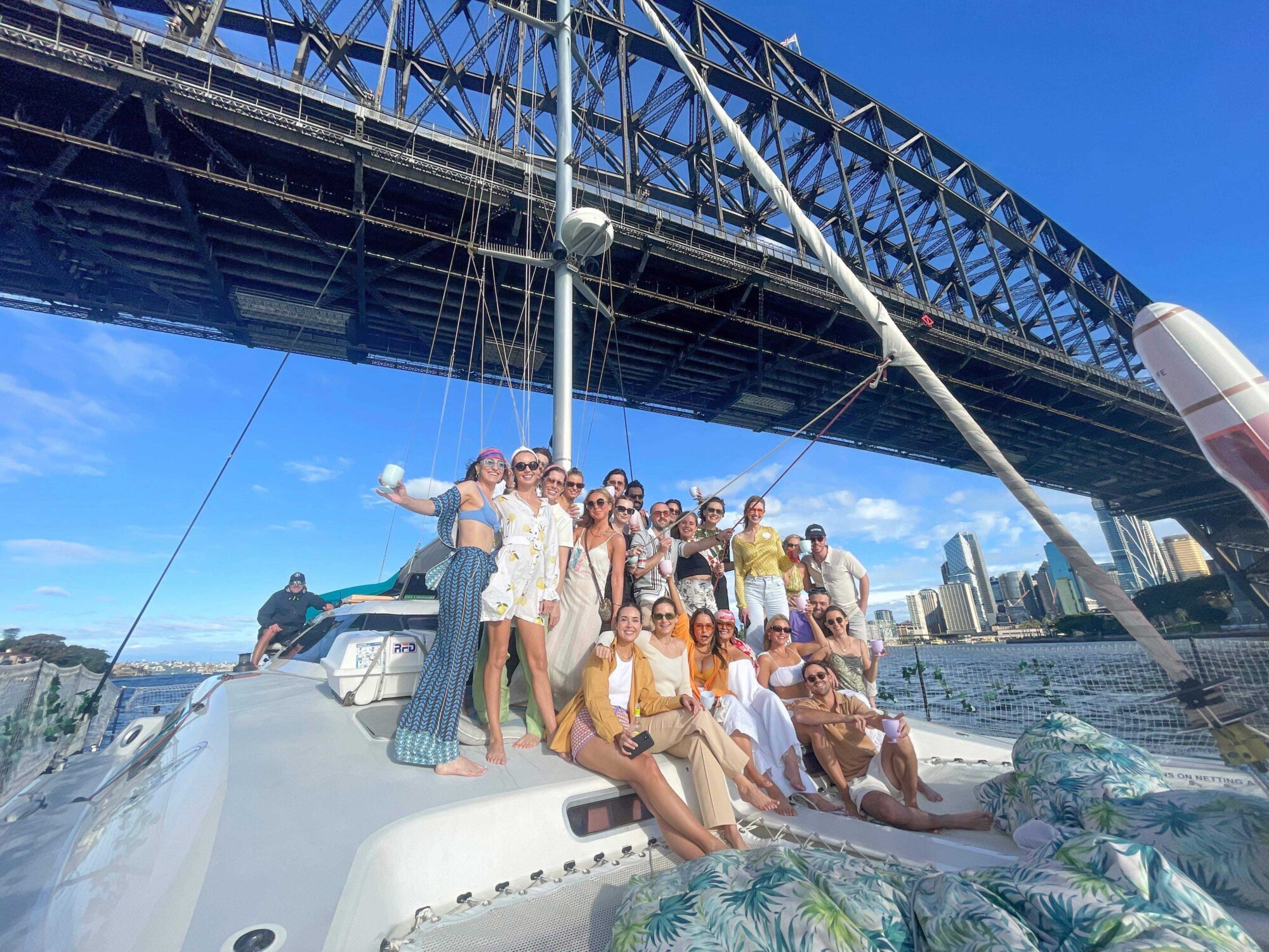 A group of people pose for a photo on a sailboat under a large bridge on a sunny day. The Yacht Social Club event features smiles and leisure as the yacht cruises past city skyscrapers in the background, showcasing an unforgettable experience.