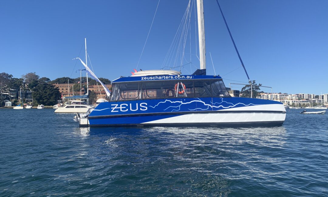 A blue and white catamaran named "Zeus" is floating on a calm body of water. The catamaran has the text "zeuscharters.com.au" on its side. Other boats and a shoreline with buildings and trees are visible in the background under a clear blue sky.