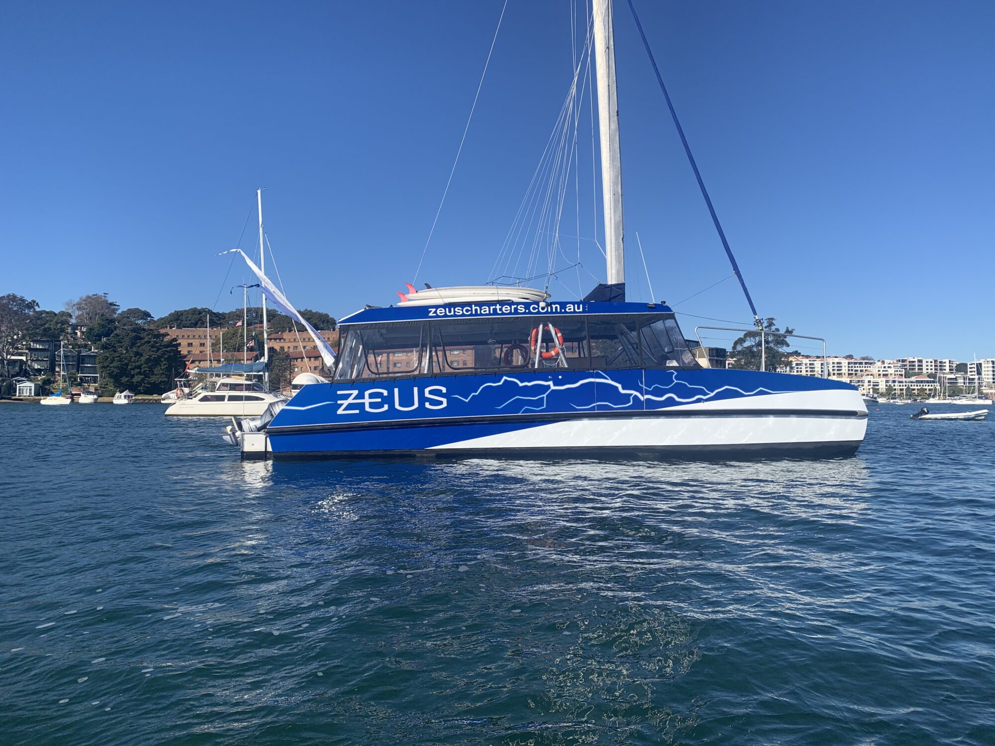 A blue and white catamaran named "Zeus" is floating on a calm body of water. The catamaran has the text "zeuscharters.com.au" on its side. Other boats and a shoreline with buildings and trees are visible in the background under a clear blue sky.