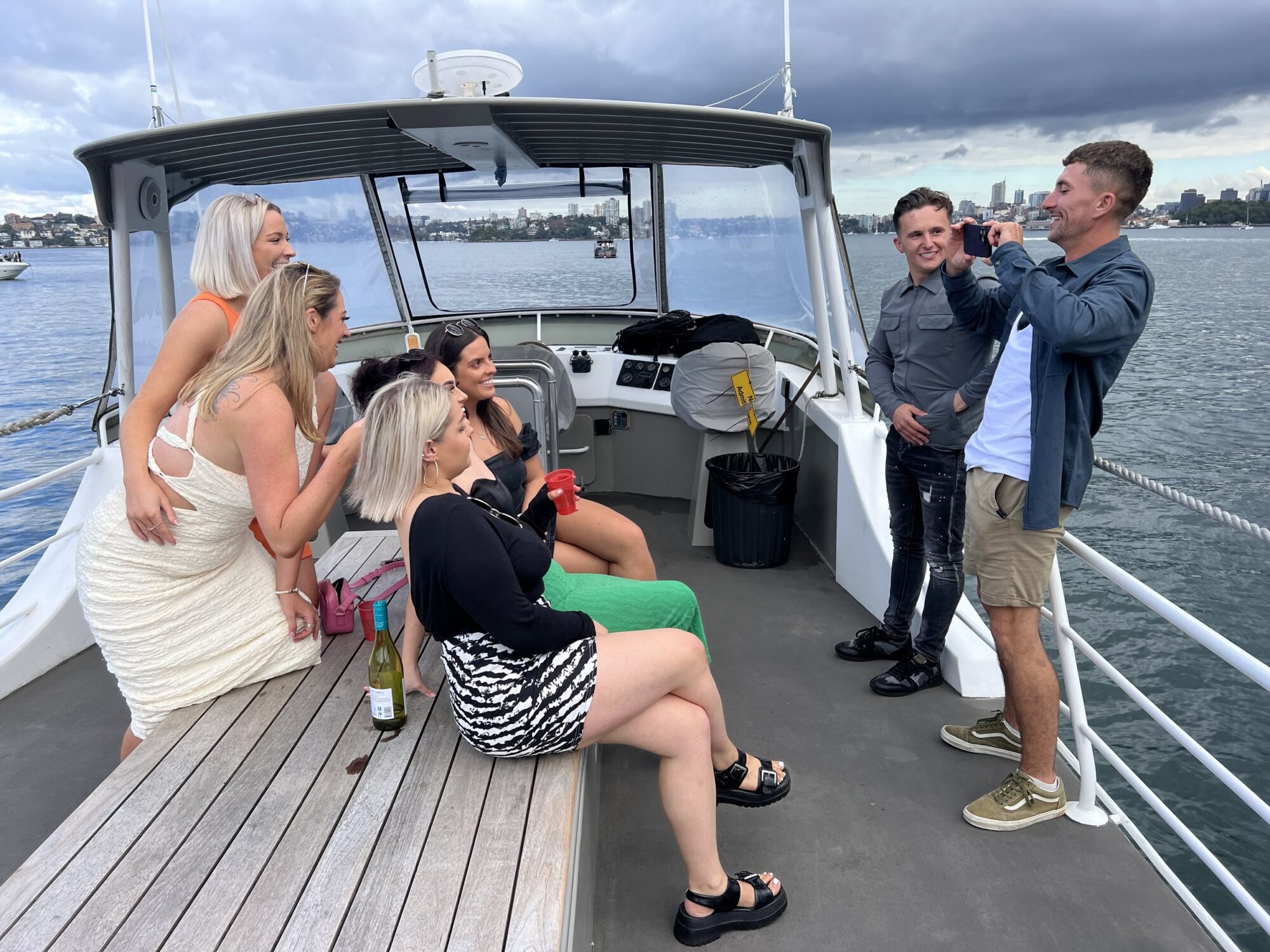 A group of six people, five women and one man, enjoy a moment on a boat deck during The Yacht Social Club Event Boat Charters. The women are sitting on a wooden bench with colorful drinks. The man stands, taking a photo of another man. The background shows a body of water and an overcast sky.