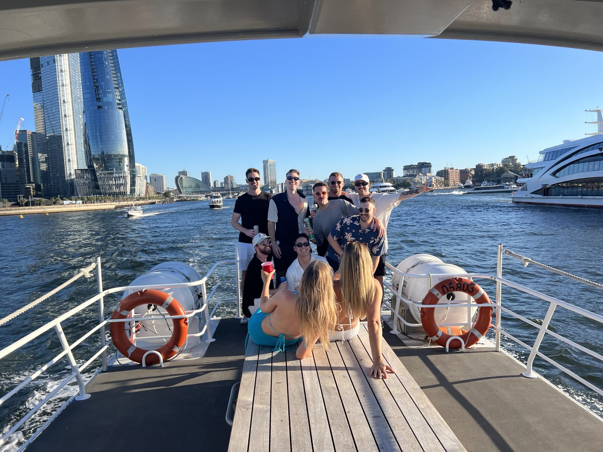 A group of nine people are posing together on the deck of a boat from Sydney Harbour Boat Hire, with skyscrapers and other boats visible in the background. The group appears to be enjoying a sunny day out on the water, with one person lounging on the deck facing the camera.