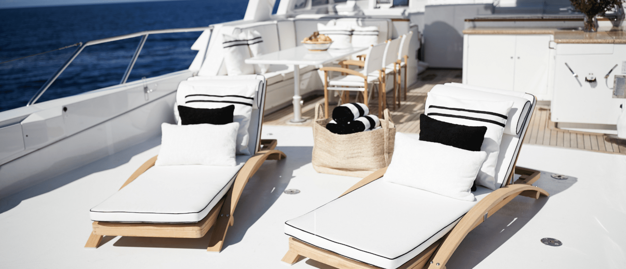 A luxurious yacht deck featuring two white cushioned sun loungers with black and white pillows, a dining area with a table and chairs in the background, and a basket filled with rolled-up towels. The deck overlooks a clear blue sea under a bright sky.