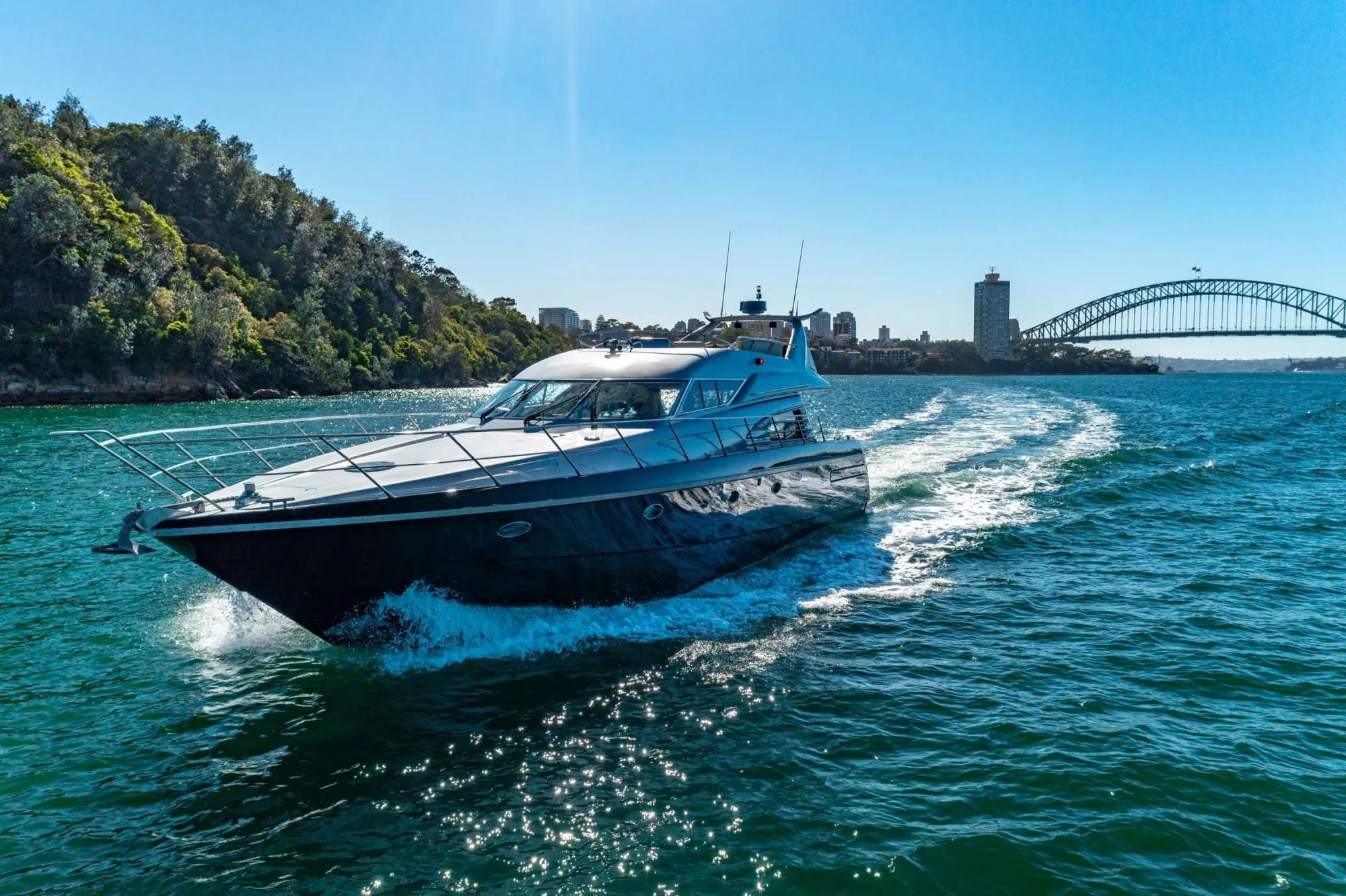 A large, sleek motor yacht cruises on clear blue waters near a lush, green coastline. The iconic Sydney Harbour Bridge is visible in the background under a bright, sunny sky. Offered by The Yacht Social Club Sydney Boat Hire, this luxury yacht leaves a trail of white waves as it moves.