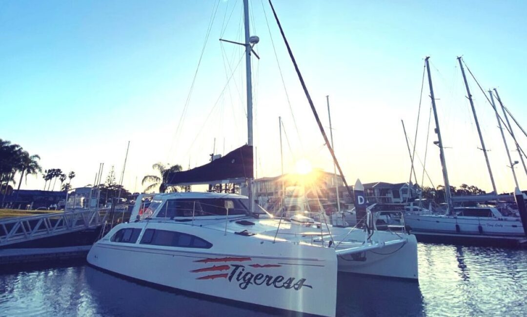 A sleek white catamaran named "Tigeress" docked at a marina during sunset. The boat has a modern design with large windows and a sail in place. The sun is low on the horizon, casting a warm glow over the calm water and nearby boats. Palm trees are visible in the background.