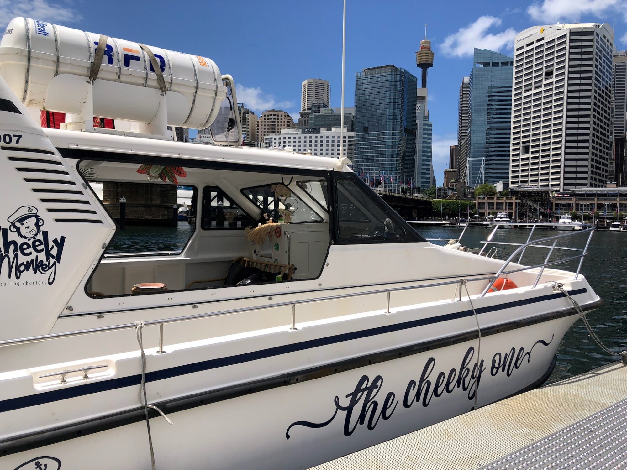 A white boat named "The Cheeky One" is docked by the water with city skyscrapers, including a tall tower, in the background. The boat has a blue-and-white life preserver on top and features a cheeky monkey cartoon on the side. Perfect for Boat Parties with The Yacht Social Club Sydney, the sky is clear and sunny.