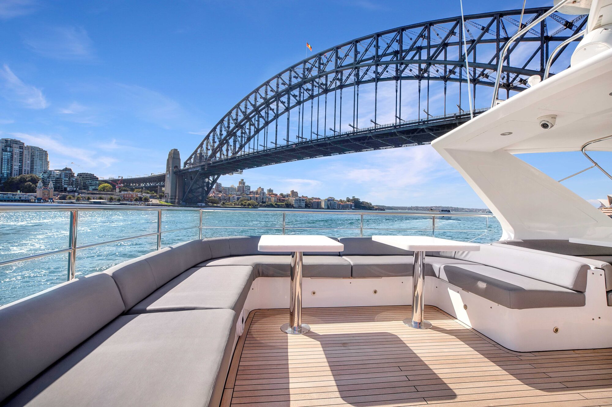 A luxurious yacht with comfortable seating and tables on its deck is cruising on a sunny day. The iconic, arched Sydney Harbour Bridge looms magnificently in the background, with the sparkling water and cityscape visible under a clear blue sky.