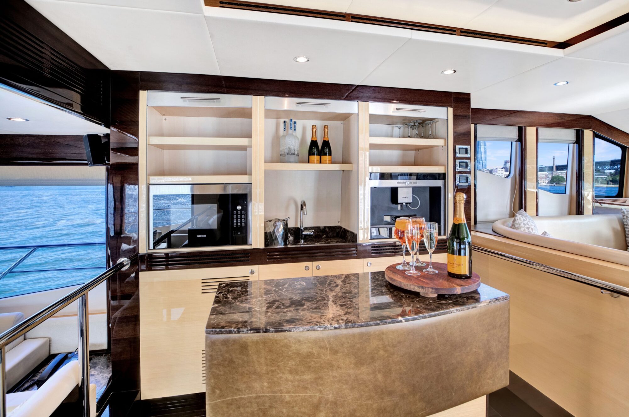 A luxurious yacht interior featuring a modern kitchen area with built-in appliances, open shelving with bottles of champagne, and a marble countertop bar with two glasses and a bottle of champagne. Large windows provide a scenic view of the ocean.