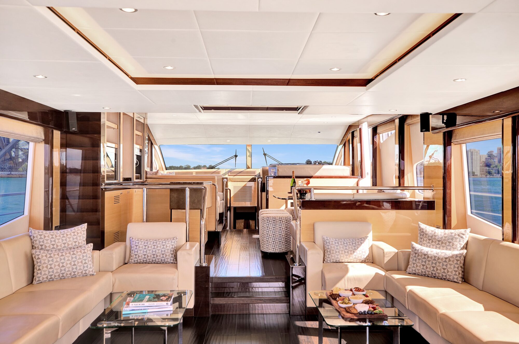 Luxury yacht interior featuring a spacious living area with cream-colored sofas, glass tables, and plush pillows. Large windows provide a panoramic view of the water, and forward of the seating area is the helm with a steering wheel and navigation equipment.