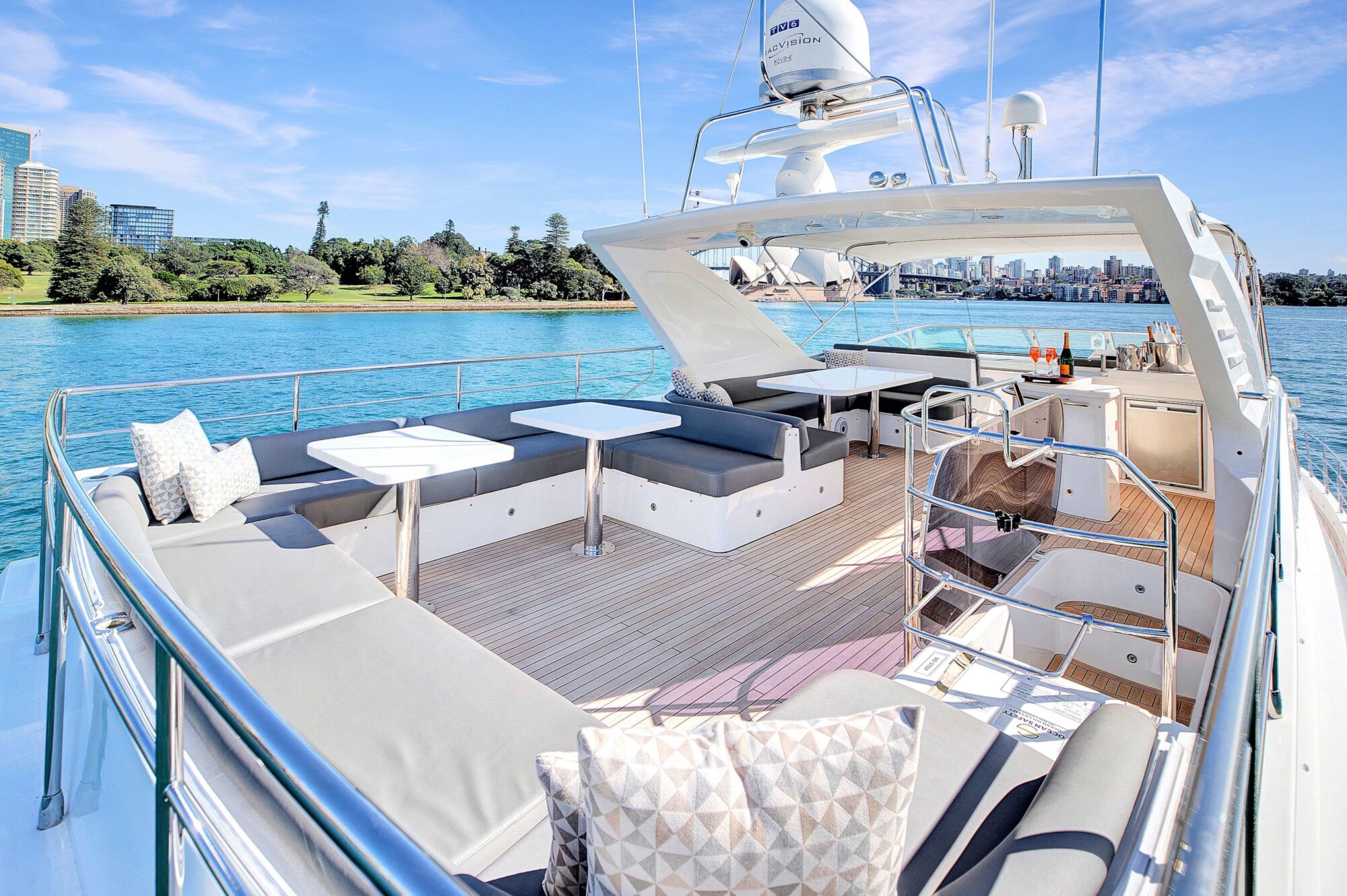 The image shows the deck of a luxury yacht with cushioned seating areas, tables, and a clear view of a waterfront cityscape. The deck is spacious, featuring sleek, modern furniture and an open-air design for relaxation with a scenic backdrop of the shoreline and buildings.