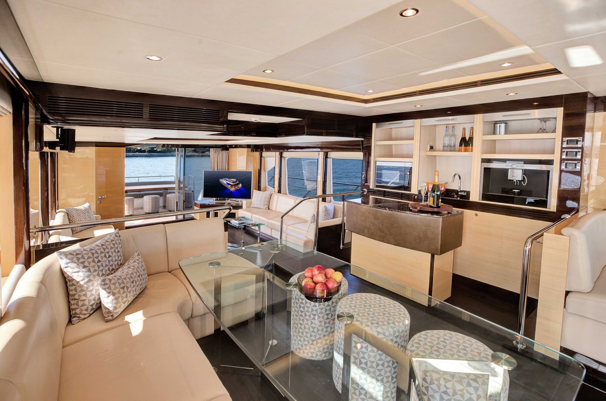 Luxurious yacht interior featuring a spacious lounge area with cream leather seating, a glass table with a bowl of red apples, modern decor, and large windows offering ocean views. The space includes a bar area with bottled drinks and a flatscreen TV.