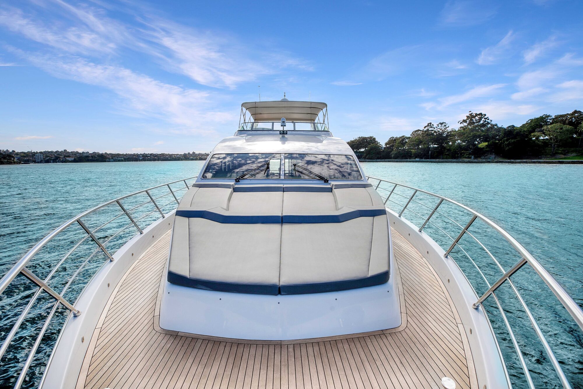 A view from the bow of a modern yacht on a sunny day, showing the boat's sleek design and expansive deck. The yacht is surrounded by calm blue waters with a shoreline and trees visible in the background under a clear blue sky with scattered clouds.