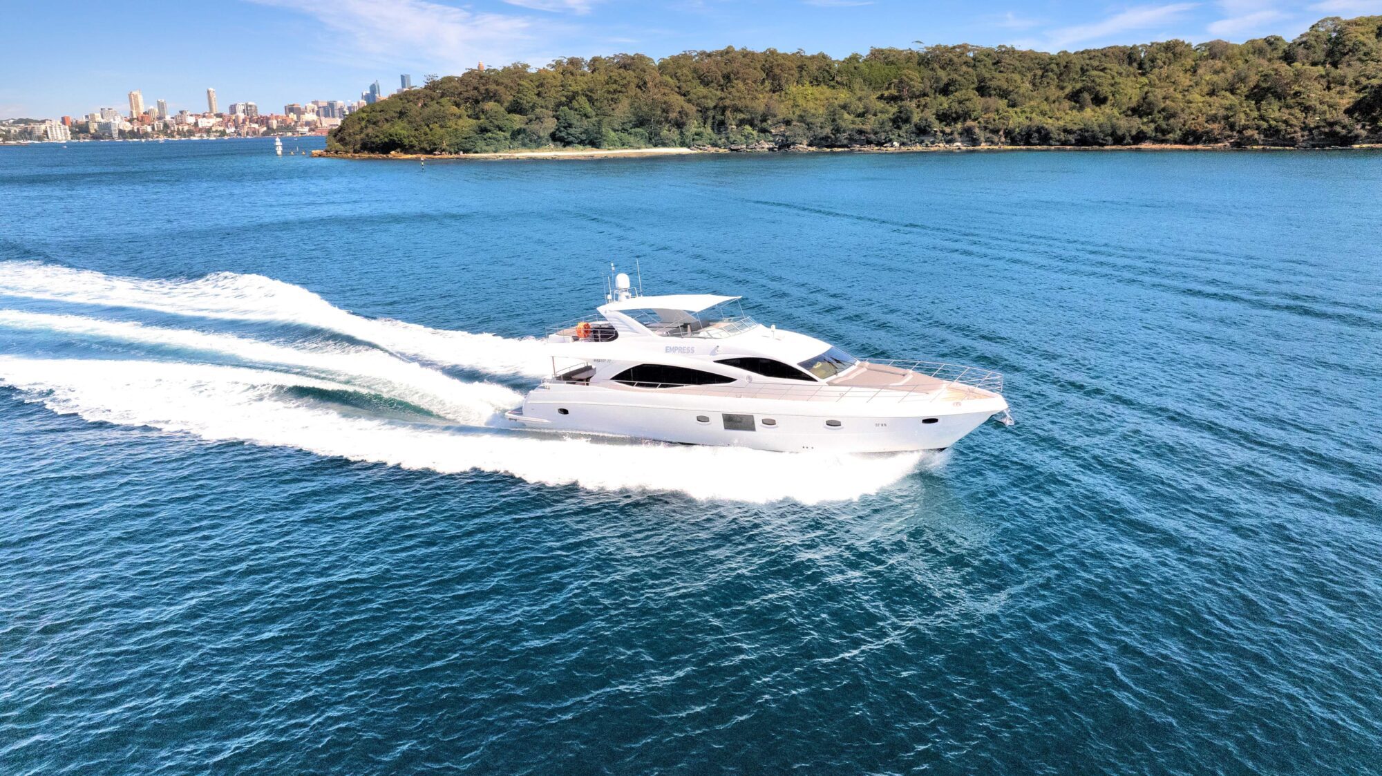 A white yacht cruises through clear blue waters near a lush, green island. The background shows a distant city skyline under a bright blue sky with a few scattered clouds, indicating a sunny day. The yacht's wake creates ripples on the water's surface.