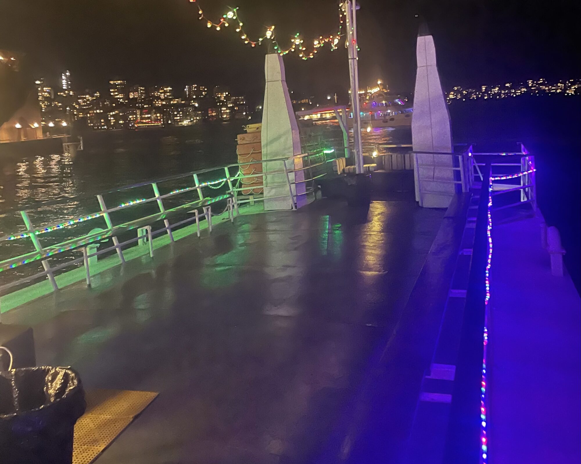 A deck of a boat is lit with colorful string lights at night. The boat is docked, and city lights are visible in the background, reflecting on the water. The atmosphere is festive, highlighted by the lights and visible structures on the deck.