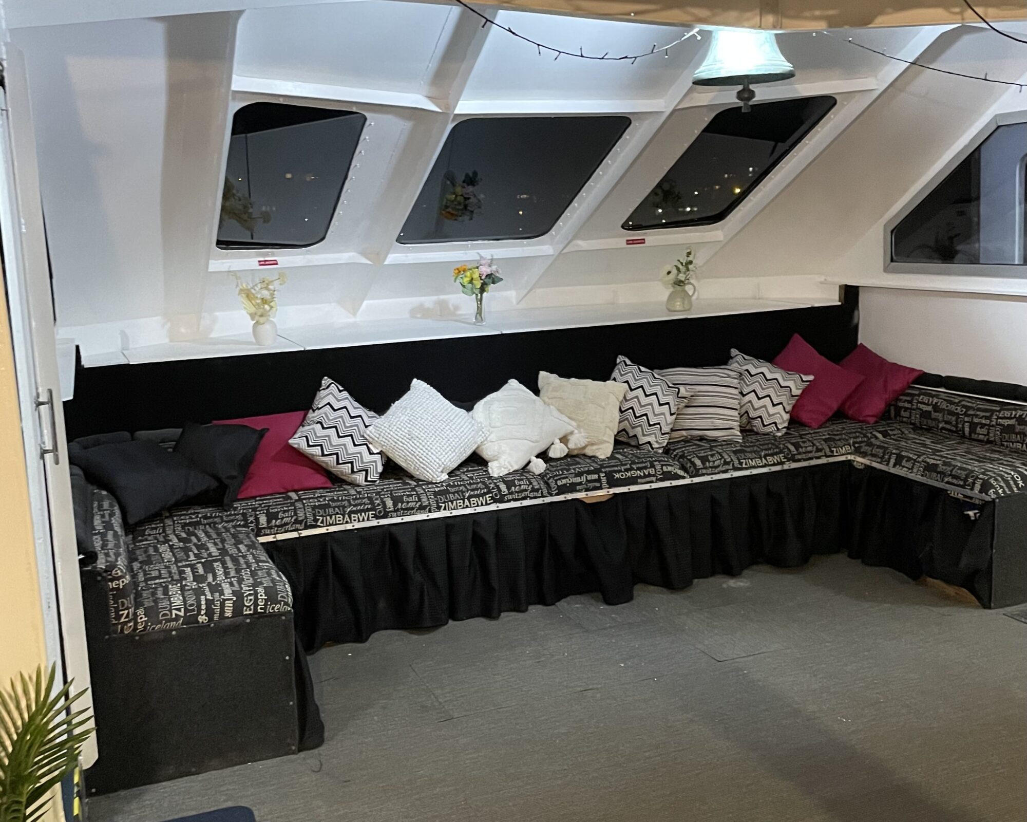 A cozy seating area with a long, L-shaped bench covered in black fabric and patterned cushions in black, white, and pink. Above the bench are three small vases with flowers positioned on a ledge in front of large windows, letting in natural light.