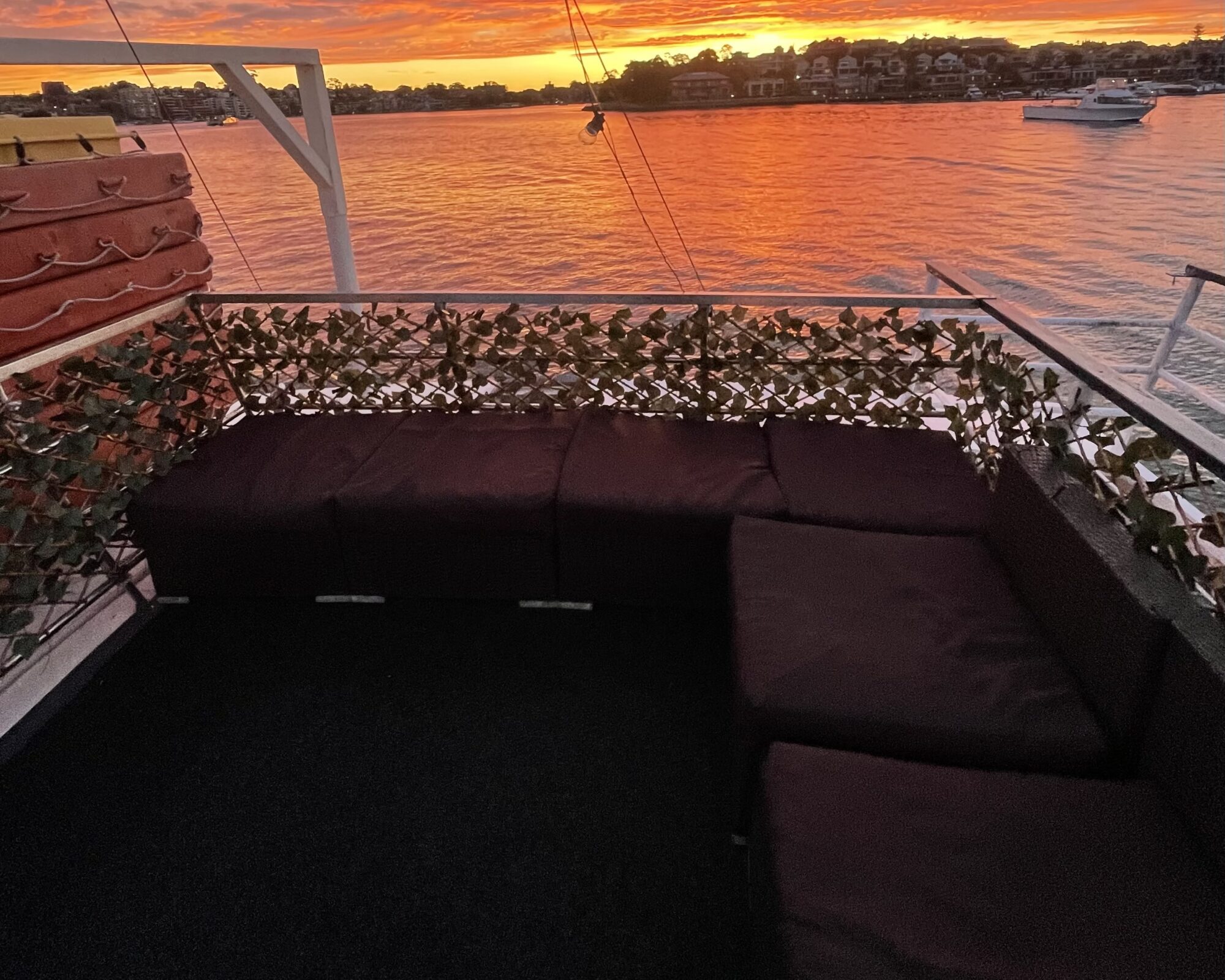 A cozy outdoor seating area with dark cushions on a boat, decorated with green foliage, overlooks a serene body of water at sunset. The sky is ablaze with vibrant orange and pink hues, with houses and a boat visible in the distance.