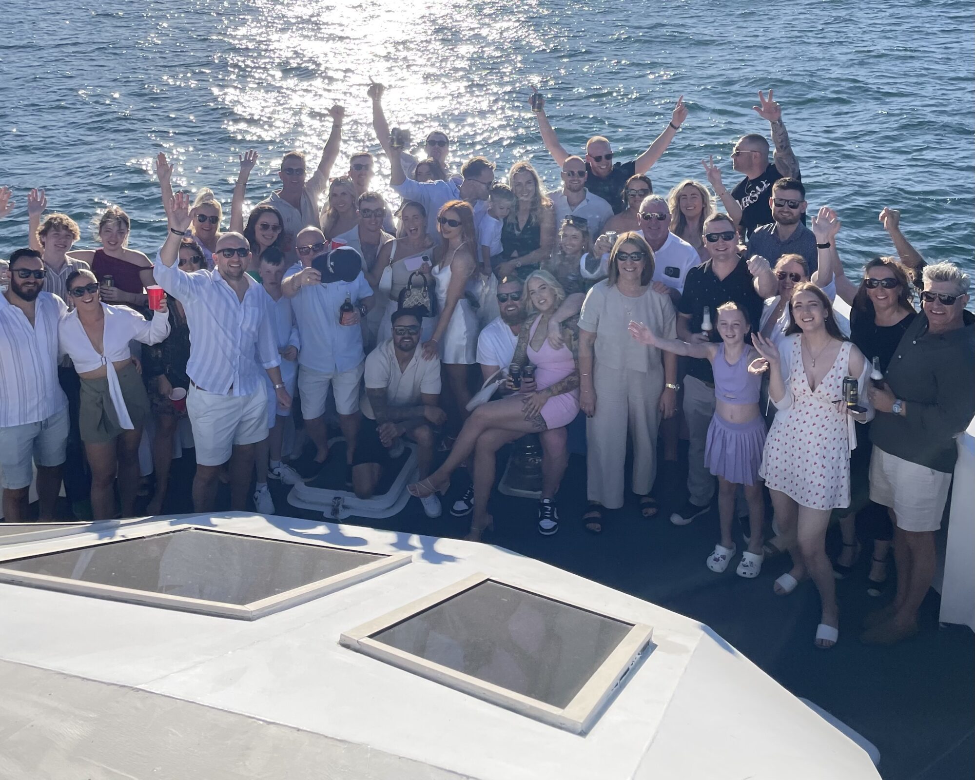 A large group of people stand together on a boat, smiling and waving their arms in the air. The sun reflects off the water in the background, casting a bright light. They seem to be enjoying a sunny day out on the water, dressed in casual summer attire.