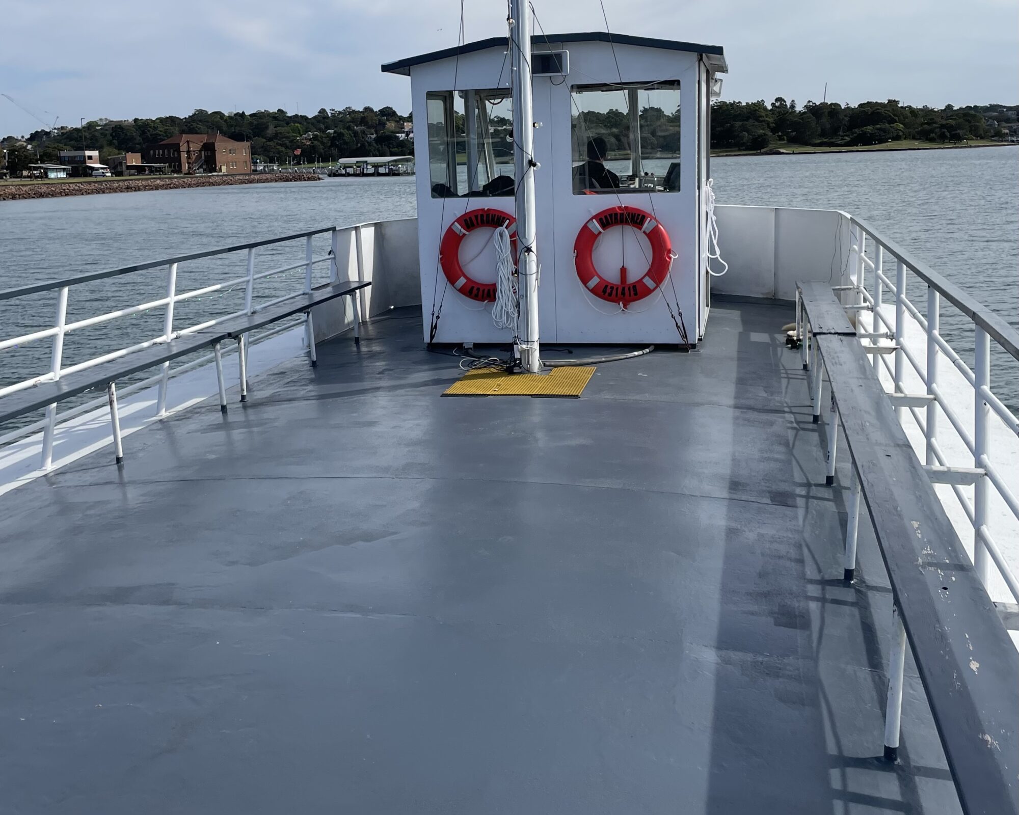 The deck of a boat with a white cabin at the center, adorned with two red life rings. The boat is sailing on a body of water, and the distant shoreline with trees and buildings is visible under a partly cloudy sky.