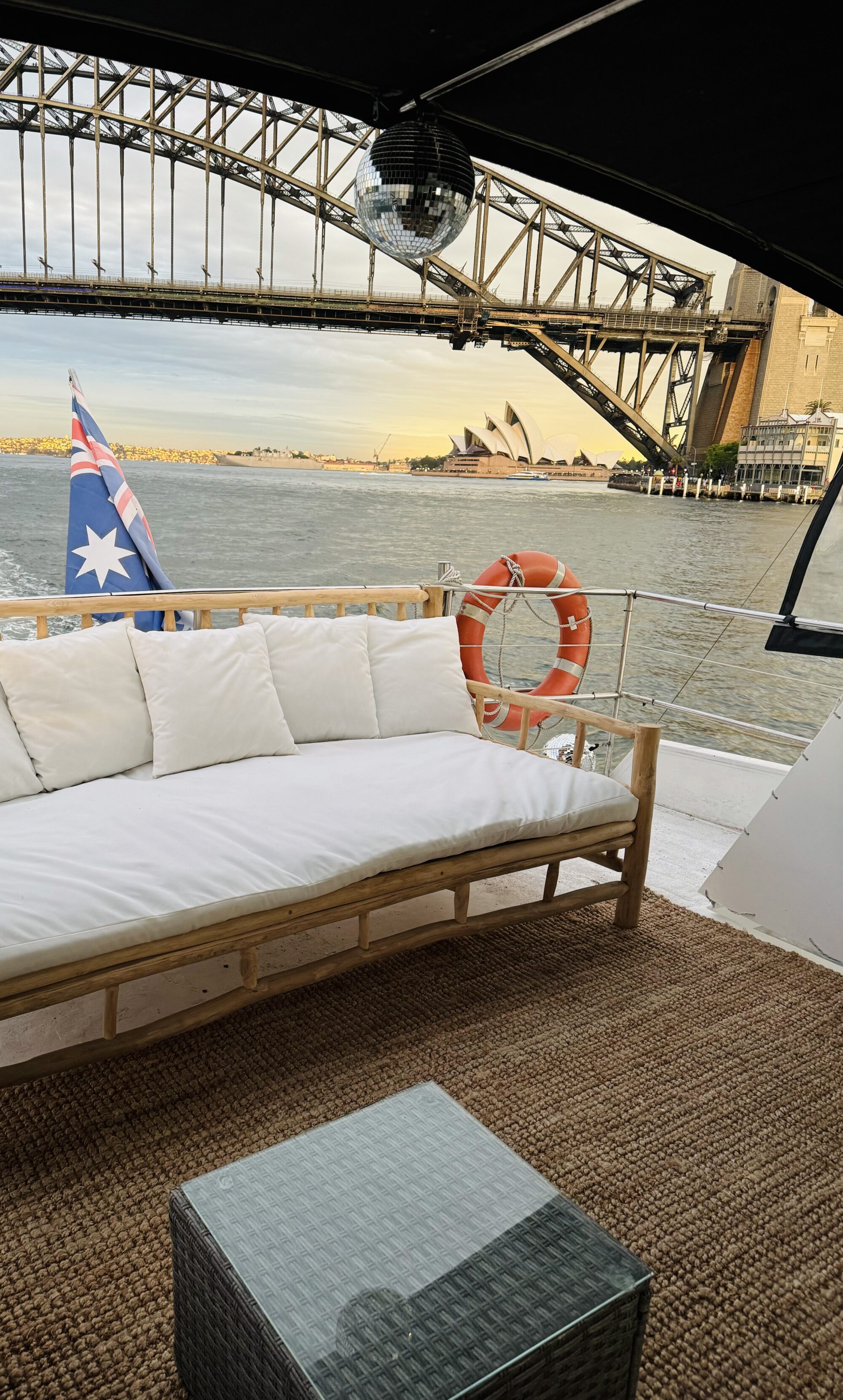 A cozy seating area on a boat, furnished with a white cushioned sofa and a glass-top table, is set against the backdrop of a bridge and the famous Sydney Opera House. An Australian flag and a life preserver are also visible on the boat.