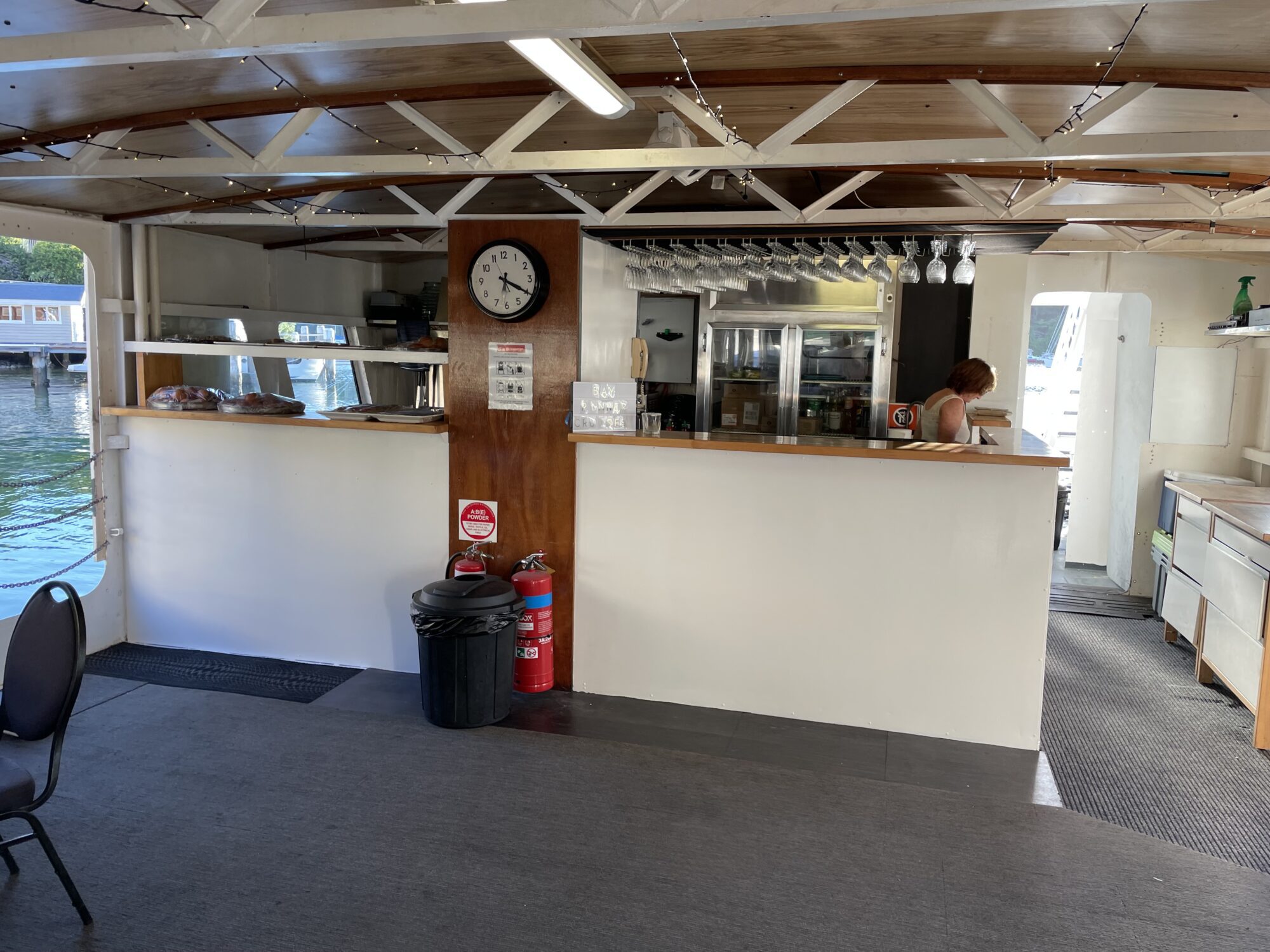 A small bar area inside a boat, with a wooden counter, a few glass racks above, and a clock on the wall. A person is seen working behind the counter. The floor is carpeted, and windows offer views of the water outside. Various safety equipment is visible.