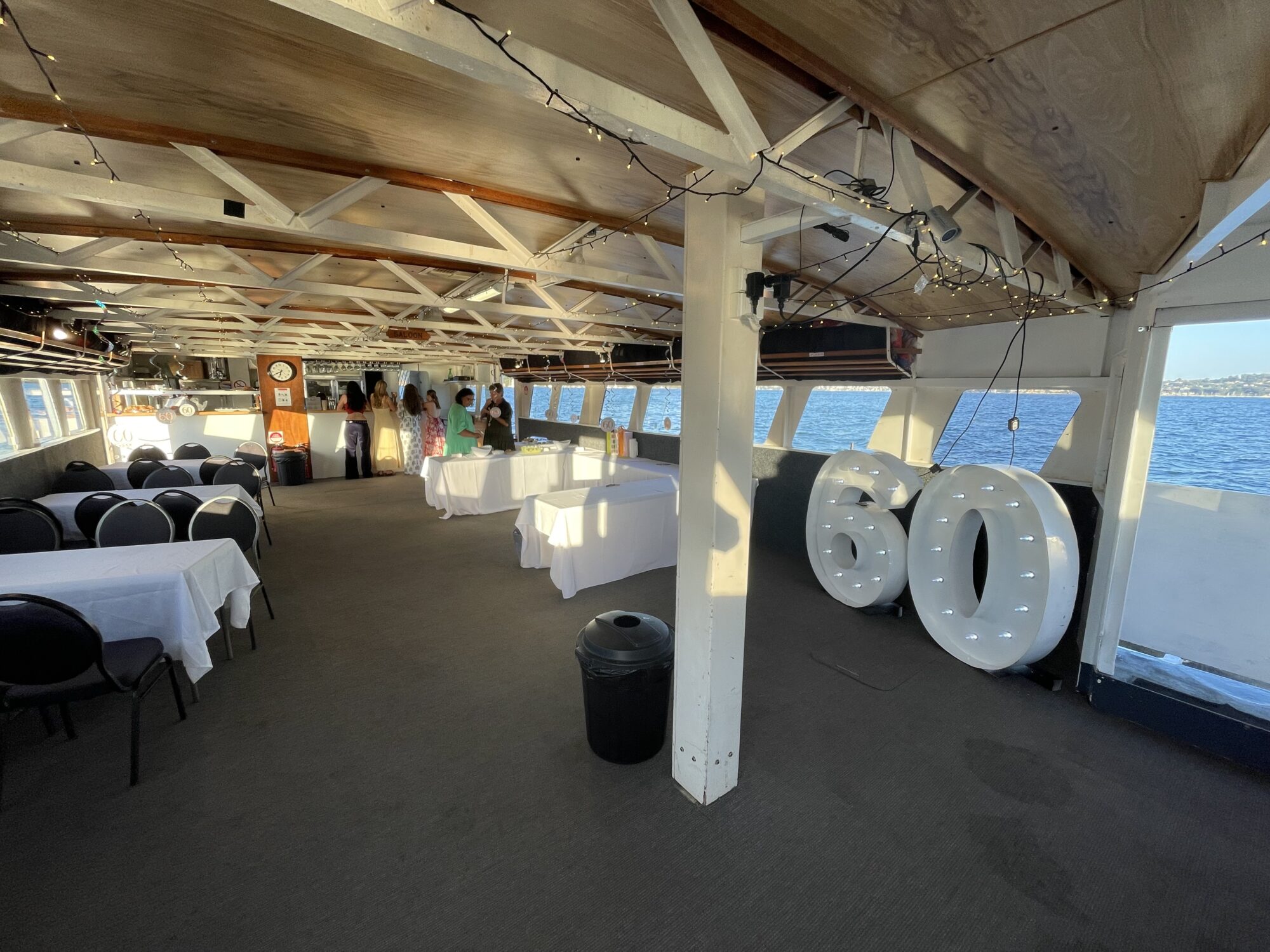 Interior view of a boat's deck set up for a celebration, featuring white tablecloth-covered tables and large balloon numbers "60". People are gathered near the back, and string lights adorn the wooden ceiling. Sunlight streams through the windows, showing the sea outside.