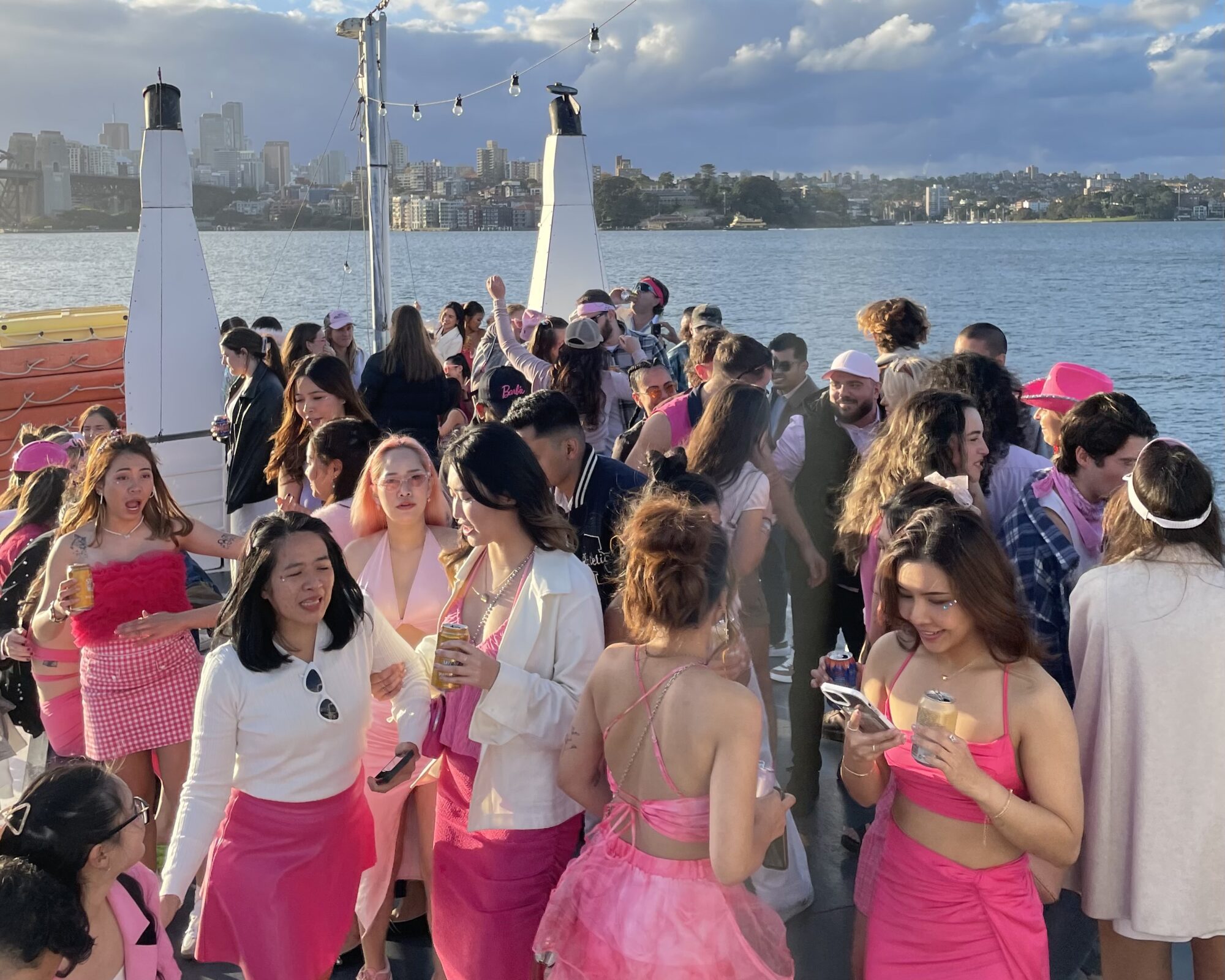 A group of people are having a lively party on a boat. The guests are dressed in pink and white attire, chatting, dancing, and taking photos. The boat is on a body of water with a city skyline and partly cloudy sky in the background.