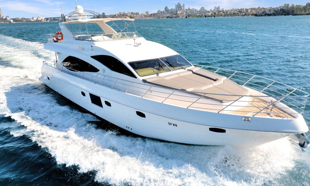 A large white luxury yacht cruising on blue waters with a city skyline in the background. The yacht has multiple decks, sleek design, and is creating a wake as it moves forward. The sun is shining, and the sky is clear.