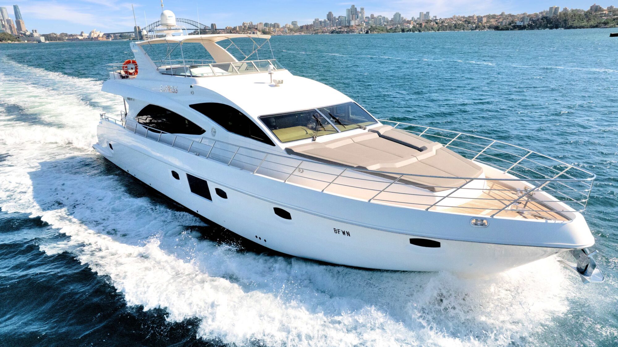 A large white luxury yacht cruising on blue waters with a city skyline in the background. The yacht has multiple decks, sleek design, and is creating a wake as it moves forward. The sun is shining, and the sky is clear.