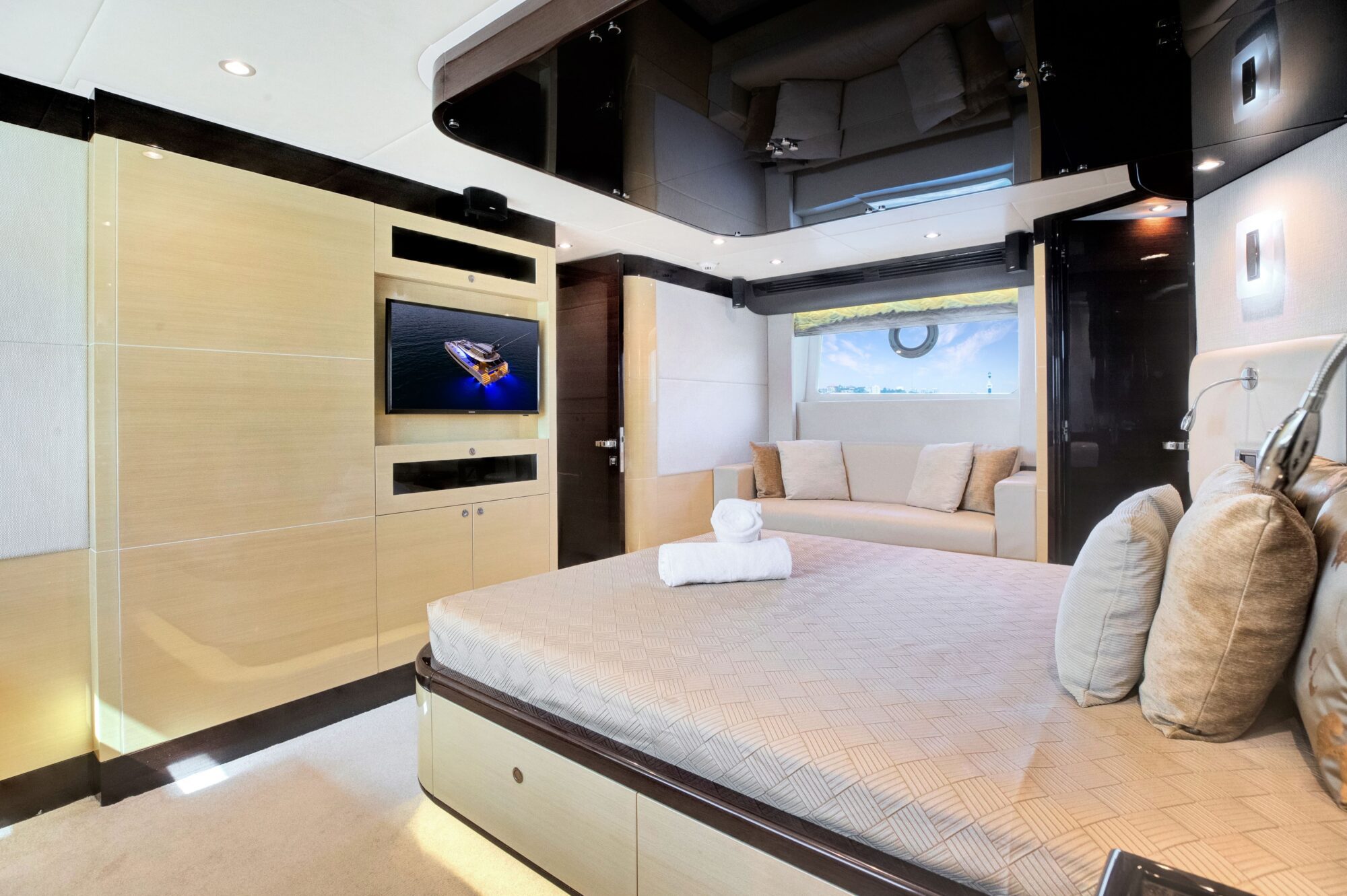 A spacious and modern yacht bedroom featuring a large bed with beige linens and multiple pillows, a flat-screen TV mounted on a light wood-paneled wall, a small sofa near a window with a view of the ocean, and soft ambient lighting creating a cozy atmosphere.