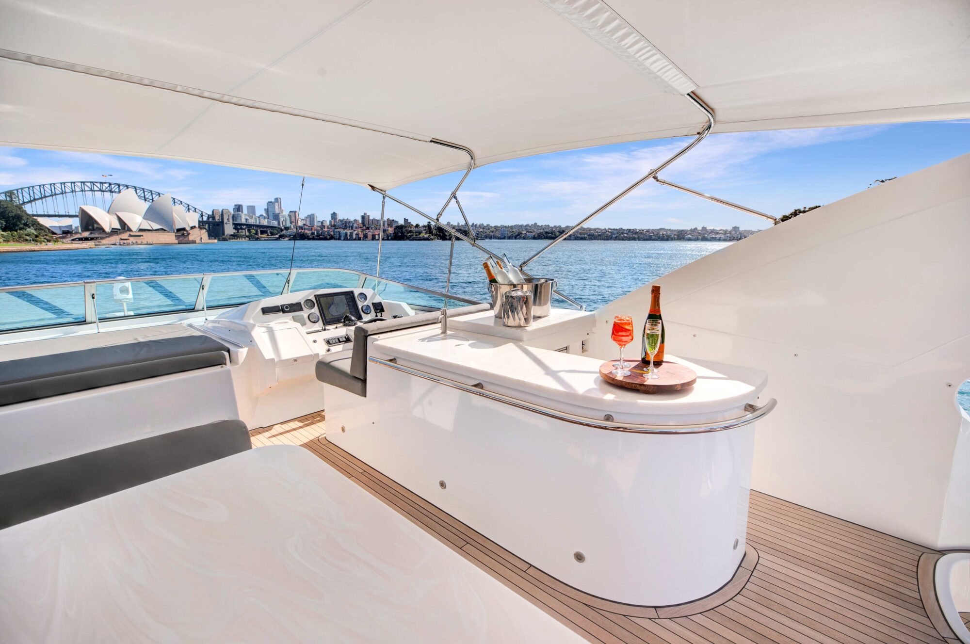 View of a yacht's upper deck featuring a bar counter with a bottle of champagne and two glasses. Behind is an urban skyline with a famous opera house and bridge over a body of water under a clear sky.