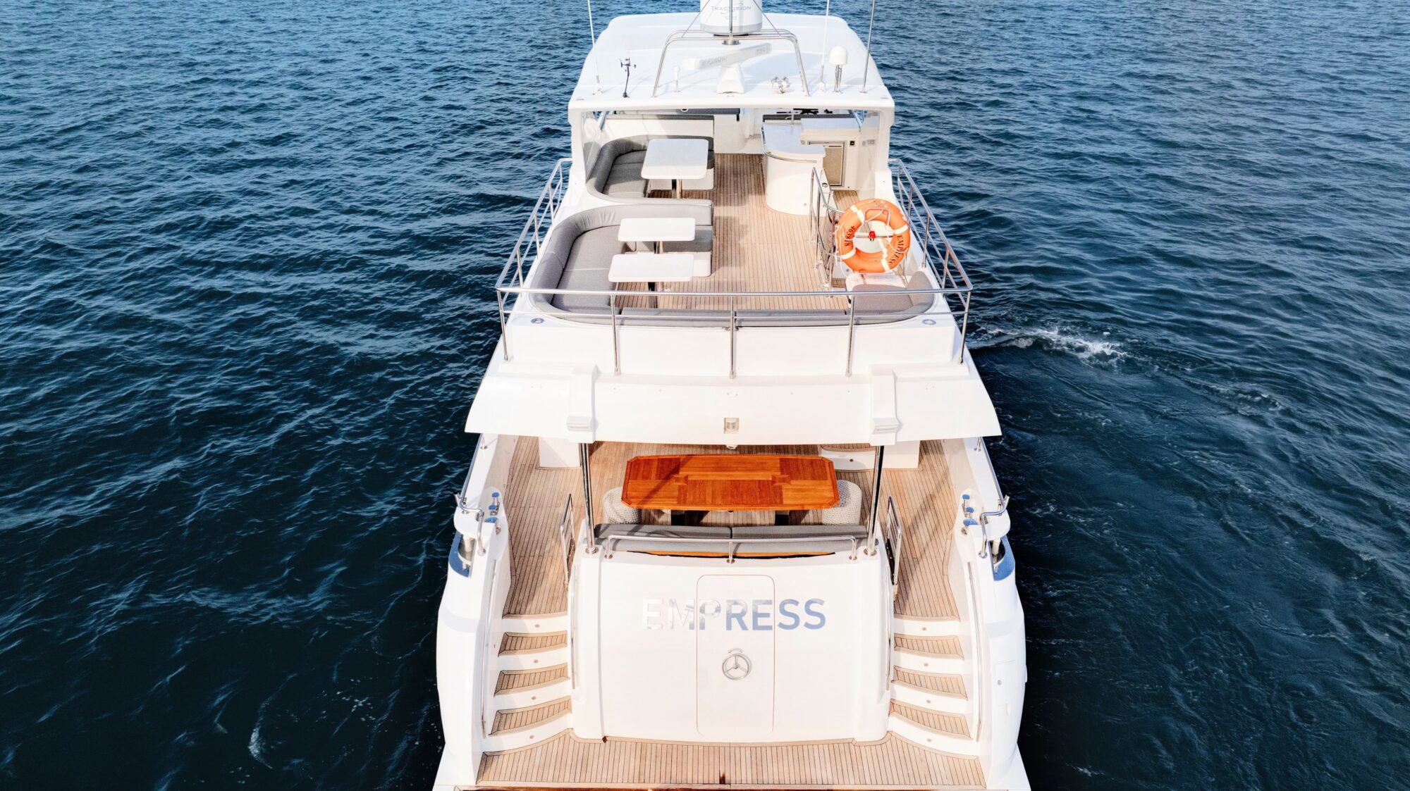 Aerial view of the back of a white luxury yacht named "EMPRESS" on the water. The yacht features multiple decks, a life ring on the upper deck, and cushioned seating areas. Two staircases lead down to the water level at the rear. The water is calm and clear.