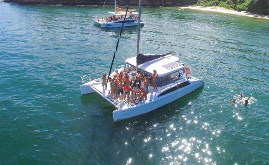 A group of people, enjoying sunny weather, gathers and parties on a large catamaran in calm, clear blue waters. Another boat and swimmers are visible in the background near a green, rocky shoreline.