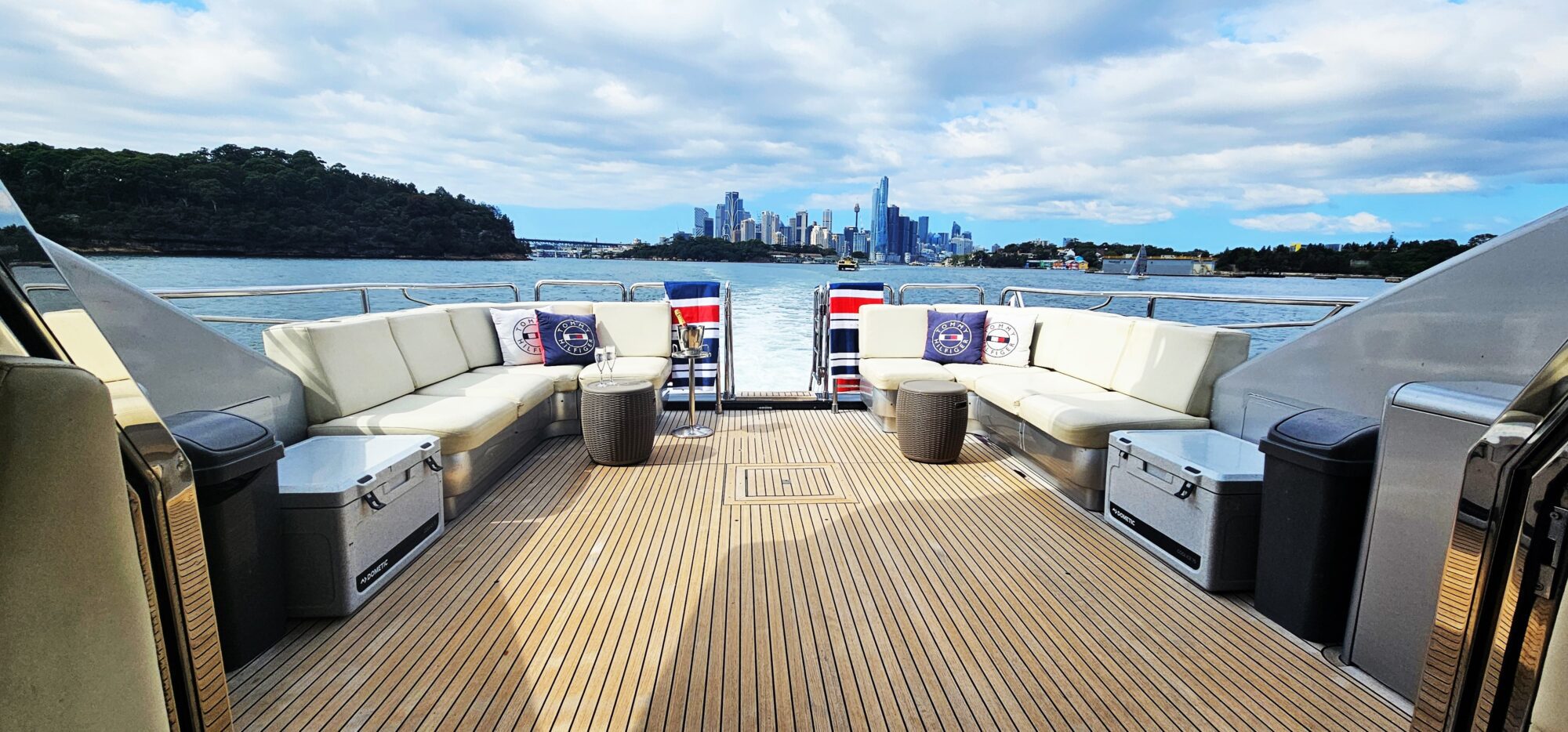 View from the deck of a luxury yacht showing white cushioned seating, modern tables, and decorated pillows. In the background, a city skyline rises under a blue sky with clouds, surrounded by water and green trees.