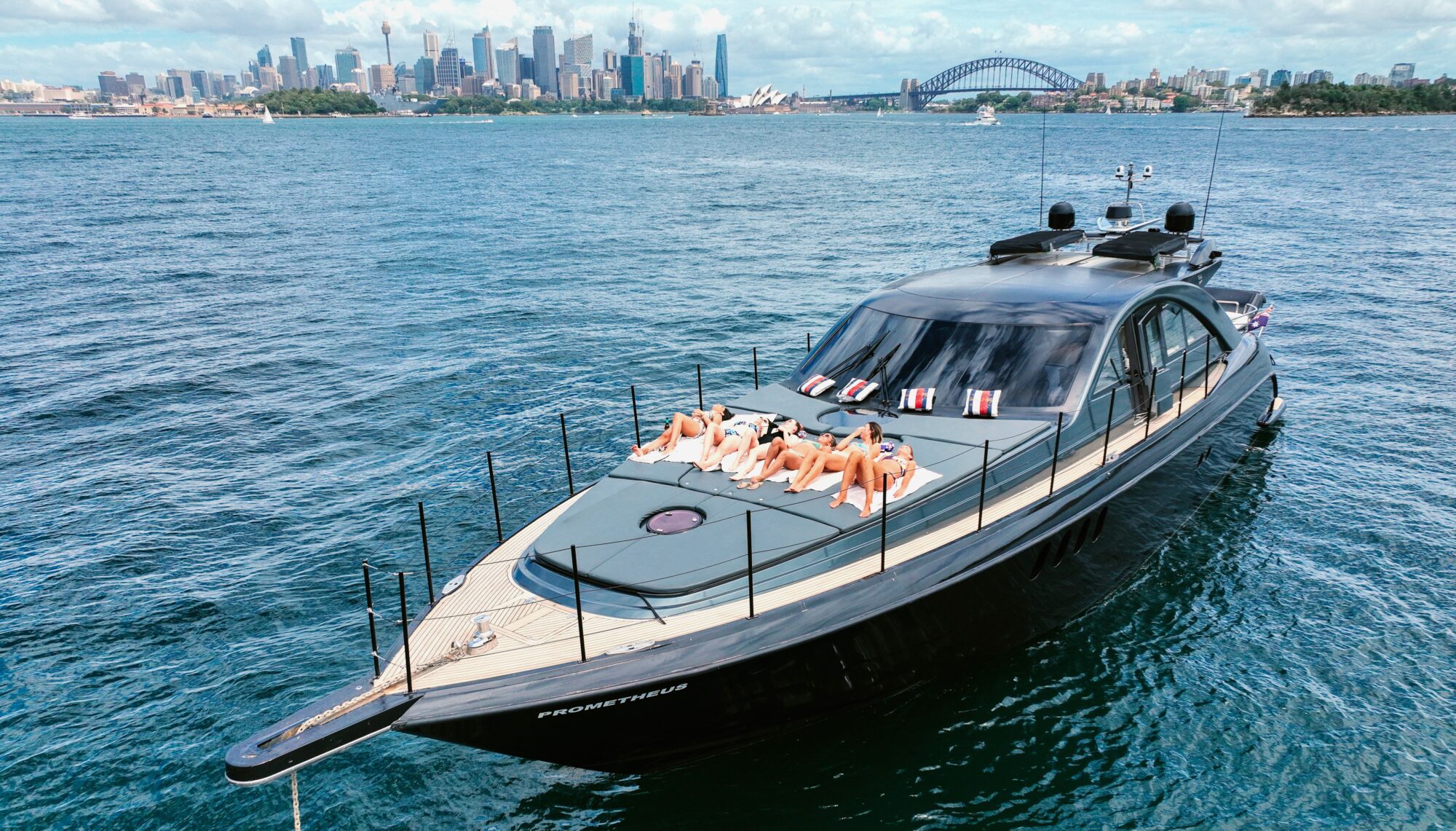 A luxury yacht with people sunbathing on its deck is anchored in a sunny harbor. The background features a city skyline with tall buildings and a prominent arched bridge over the water.