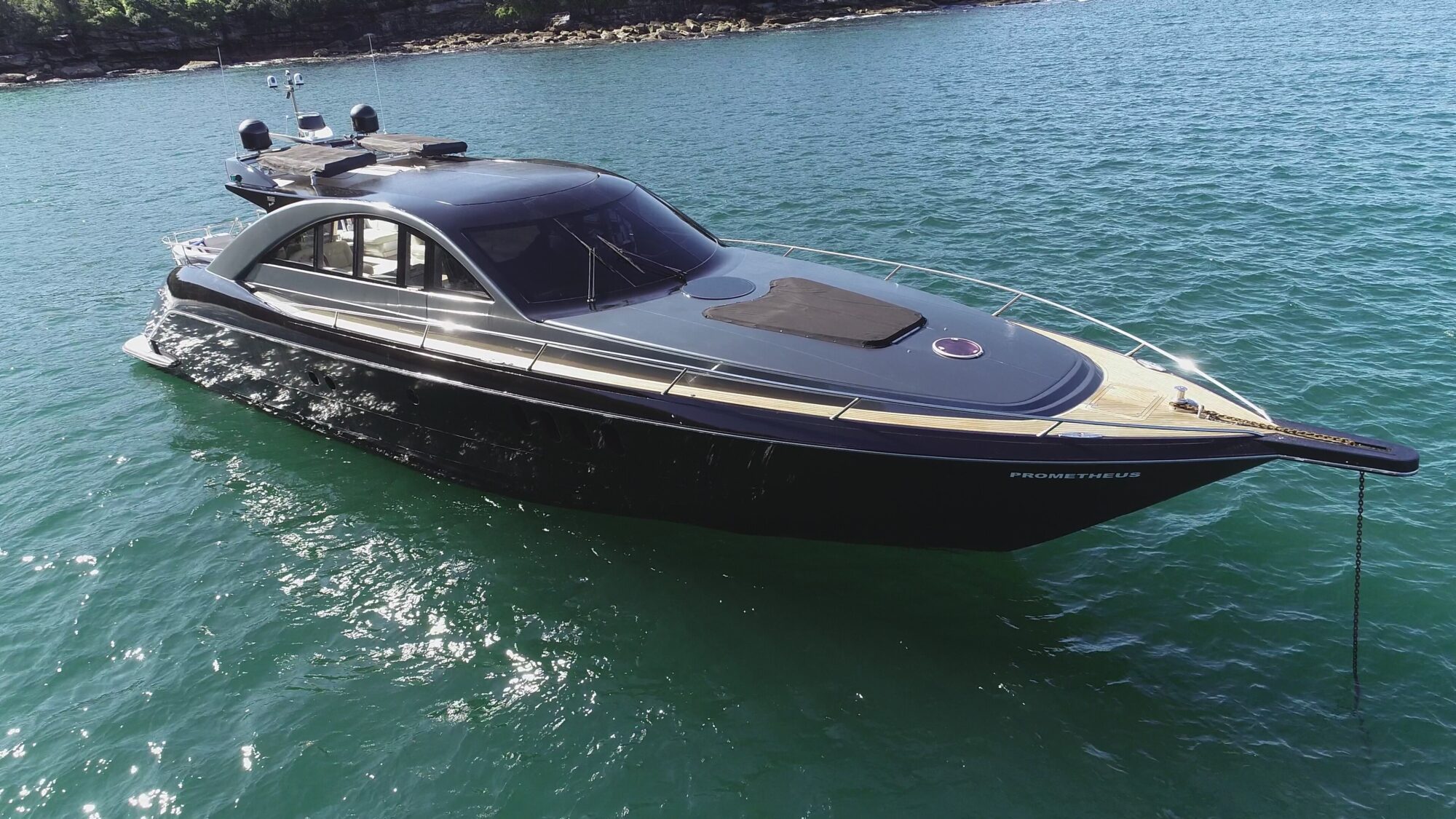 A sleek black luxury yacht named "PROMETHEUS" is anchored on calm, turquoise waters. The vessel has a modern design with tinted windows, and a small area of rocky shoreline is visible in the background.