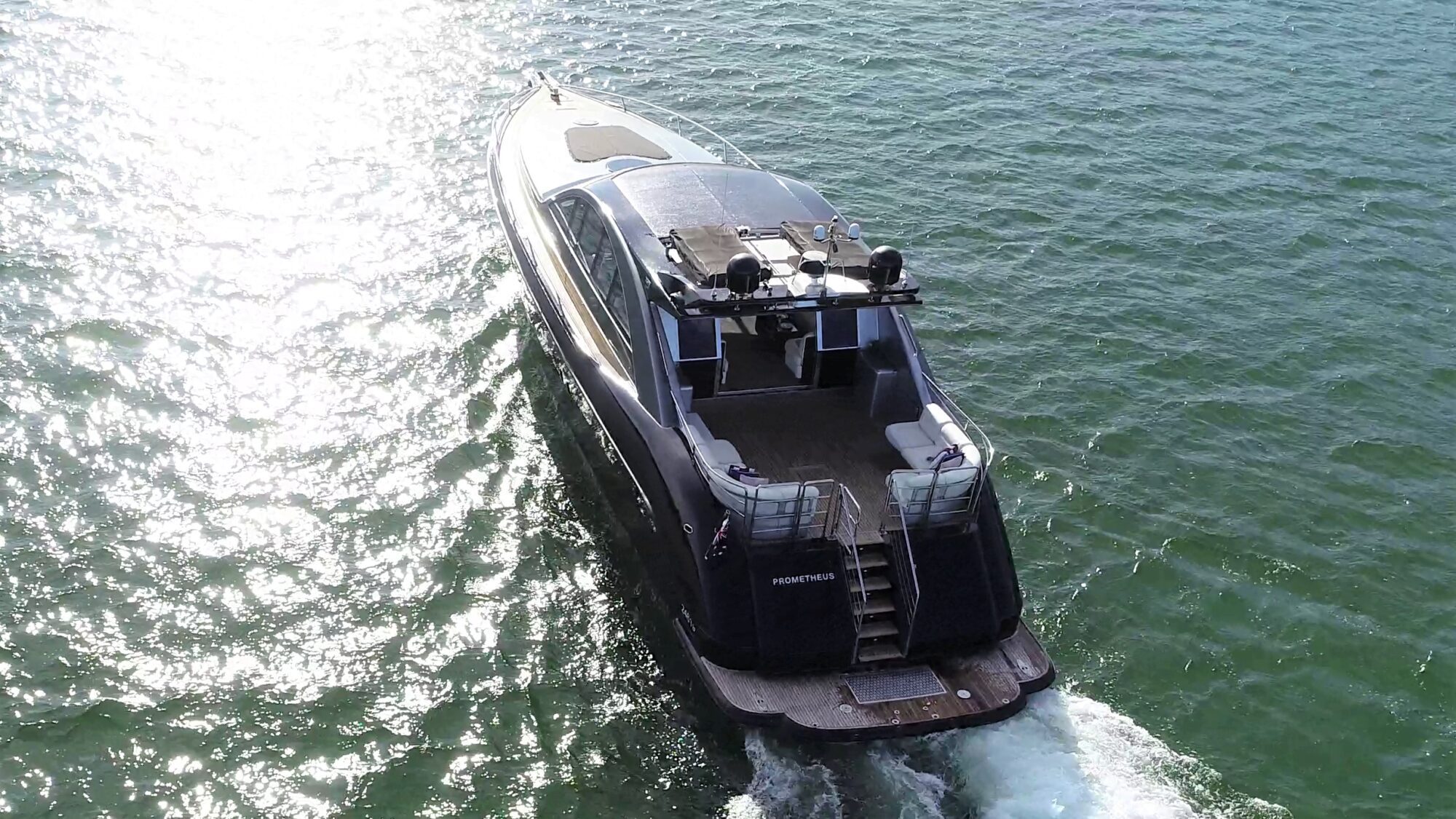 A luxury motor yacht cruising through clear, sunlit water. The vessel has a sleek design with visible deck space at the rear and a wake forming behind it as it moves.
