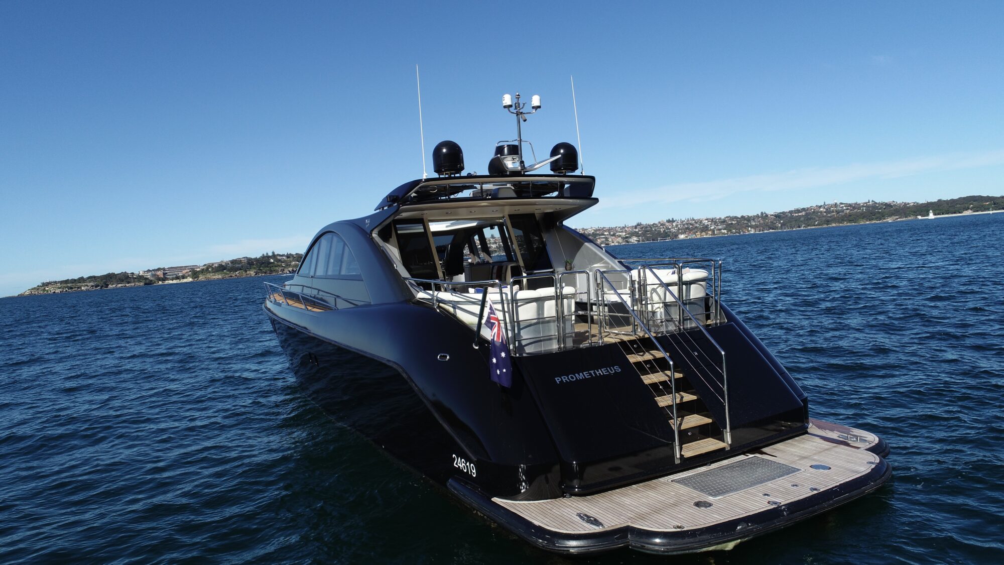 A sleek black yacht named "Prometheus" sails on a calm blue sea under a clear sky. The yacht features a spacious deck and modern design. Coastline and distant buildings are visible on the horizon.