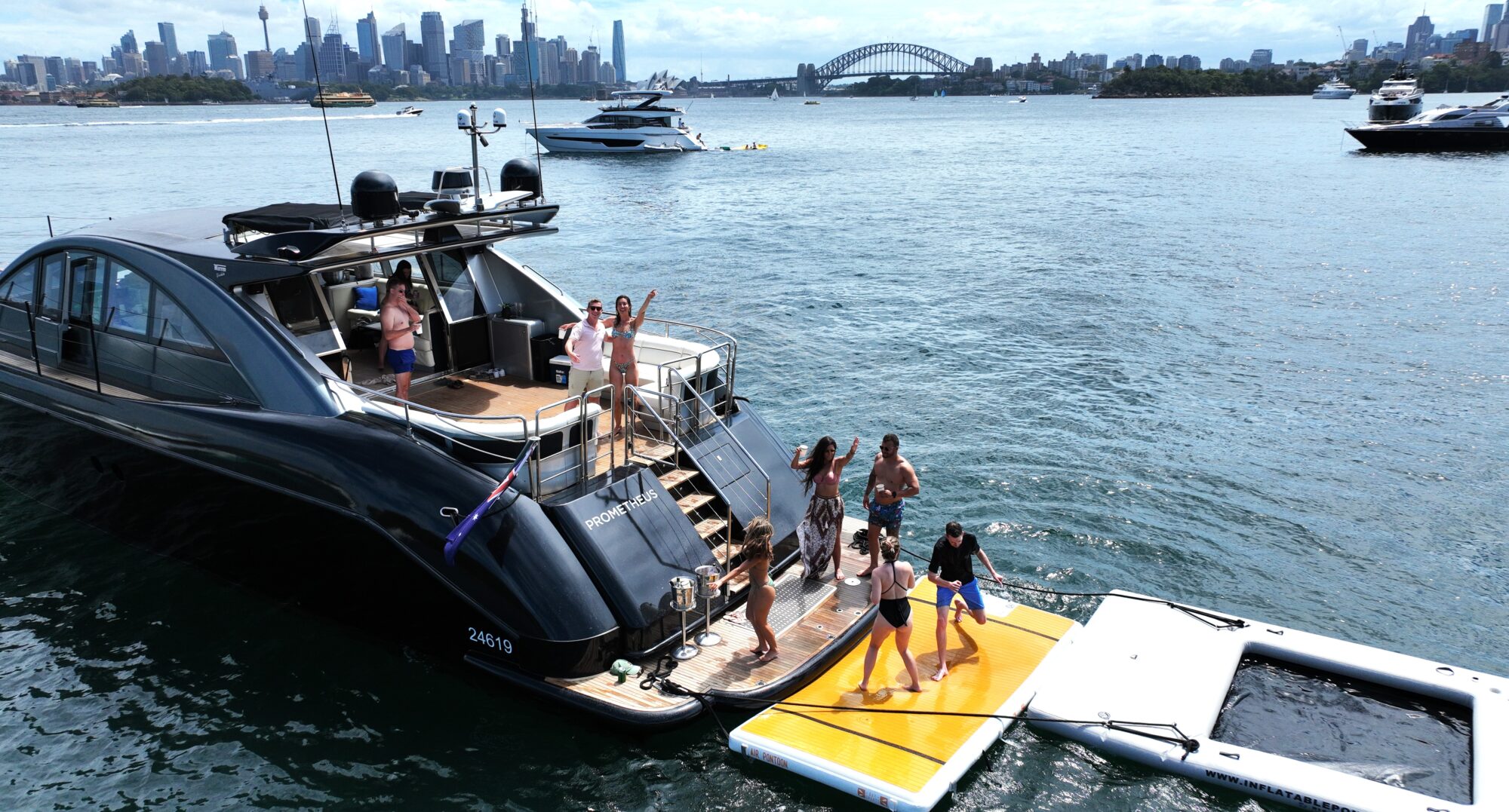 A group of people are enjoying a sunny day on a luxury yacht in the harbor. Some are on the boat’s deck, while others are on a floating platform. The city skyline and a large bridge are visible in the background.