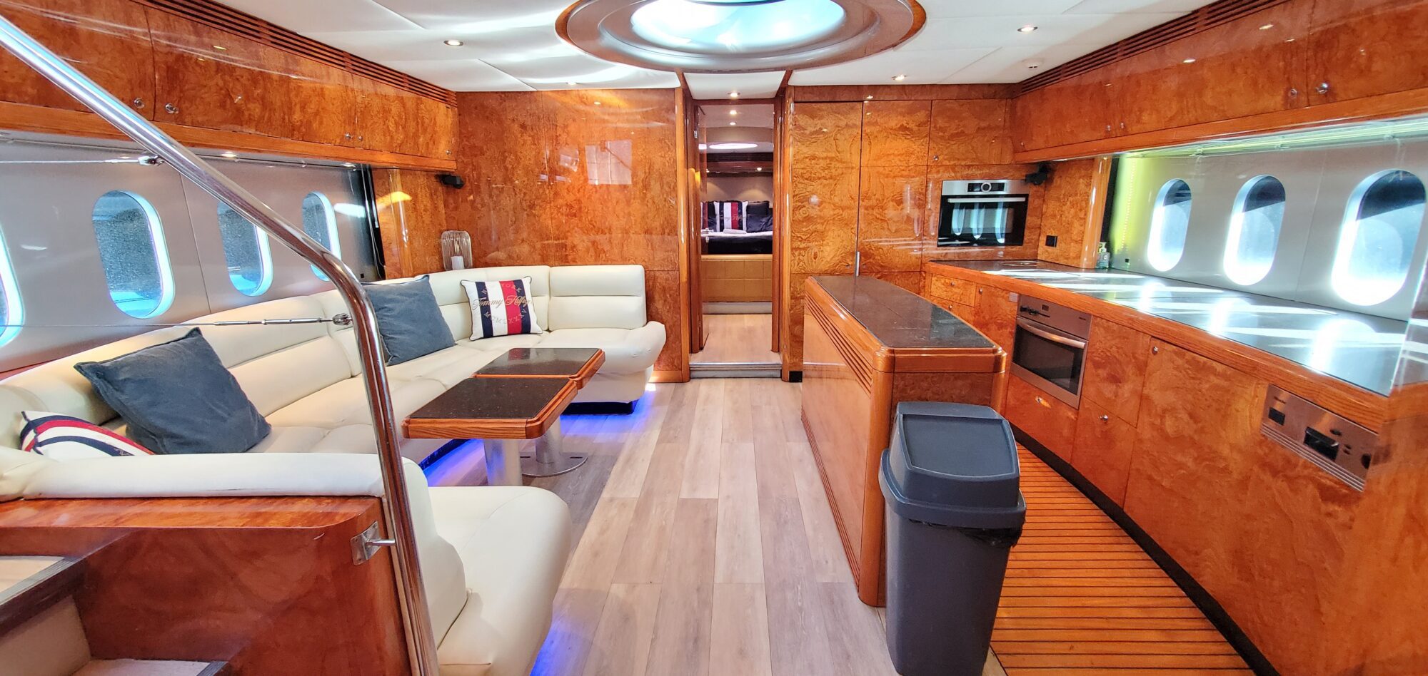 Luxurious yacht interior with wooden paneling and a modern design. Features a cream-colored seating area with decorative pillows, a sleek kitchen with stainless steel appliances, and ambient lighting. Porthole windows line the walls, offering a nautical view.