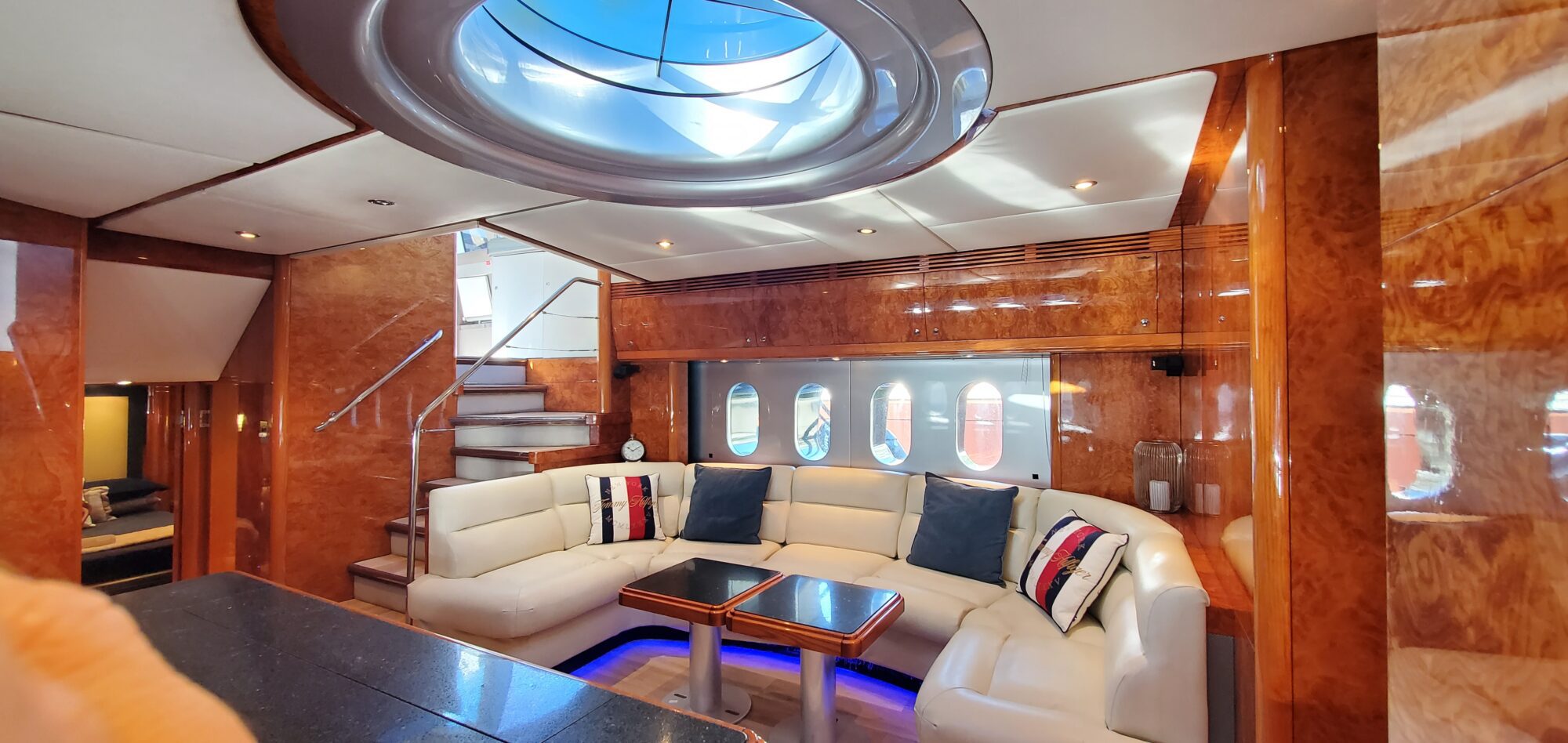Luxurious yacht interior with a round skylight, plush white seating, and a wooden finish. Features a small table with throw pillows on the couch and porthole-style windows. Elegant lighting adds a warm ambiance.