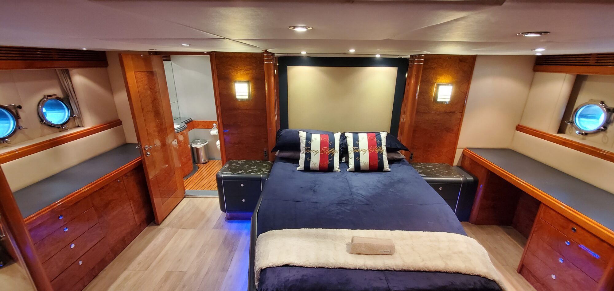 Luxurious yacht bedroom featuring a central bed with a dark blue cover and striped pillows. The room has warm wooden accents, two portholes, soft lighting, and an en-suite bathroom with modern fixtures.