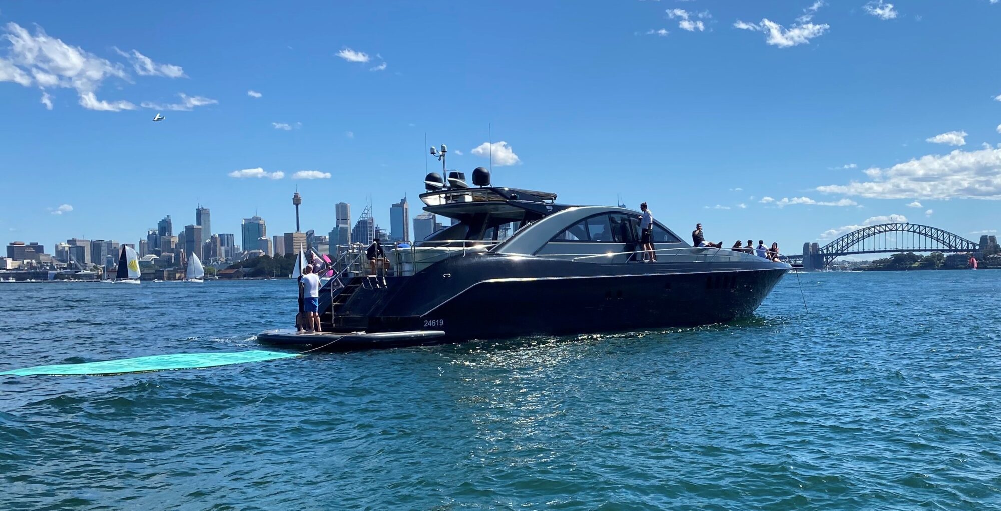 A sleek black yacht is cruising on the water with several people on board. The Sydney skyline, including the Opera House and Harbor Bridge, is visible in the background under a clear blue sky.