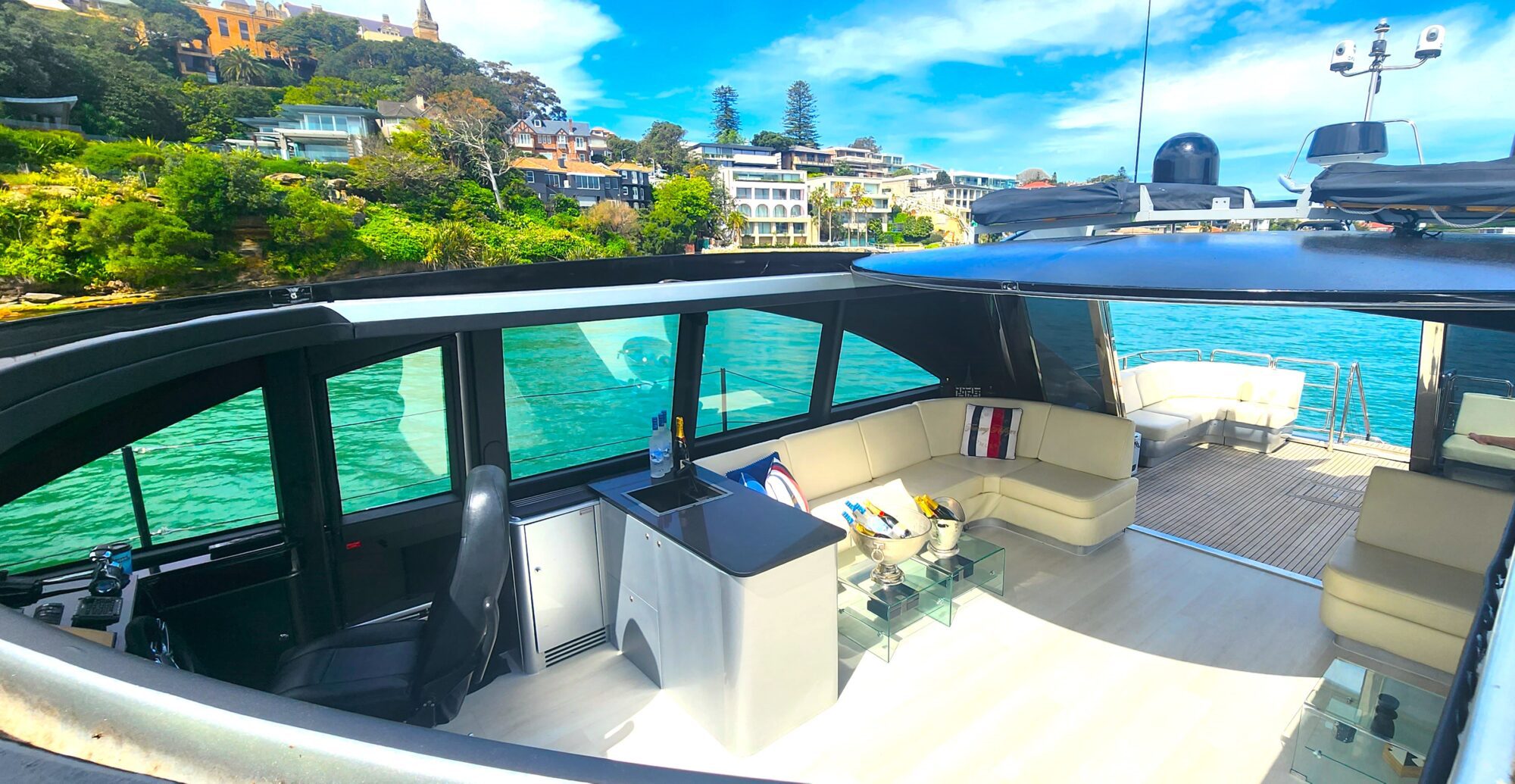 A luxurious yacht interior with cream seating, a small bar with bottles, and glass tables. The yacht is on turquoise water near a lush green coastline with elegant houses under a bright blue sky.