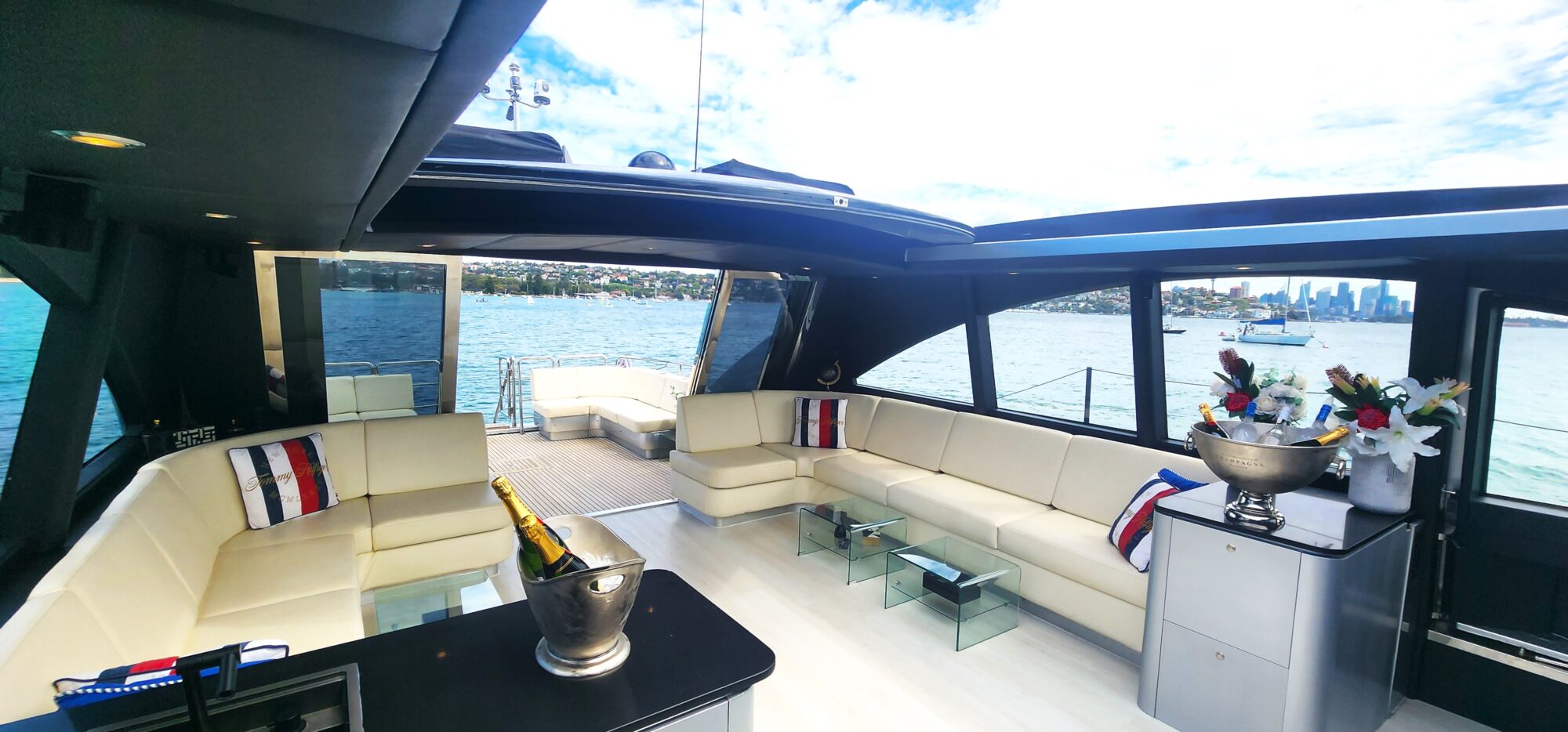 Luxurious yacht interior featuring cream leather seating with striped cushions, a black table with champagne bottles, and floral arrangements. Large windows provide views of a bay and distant city skyline under a partly cloudy sky.