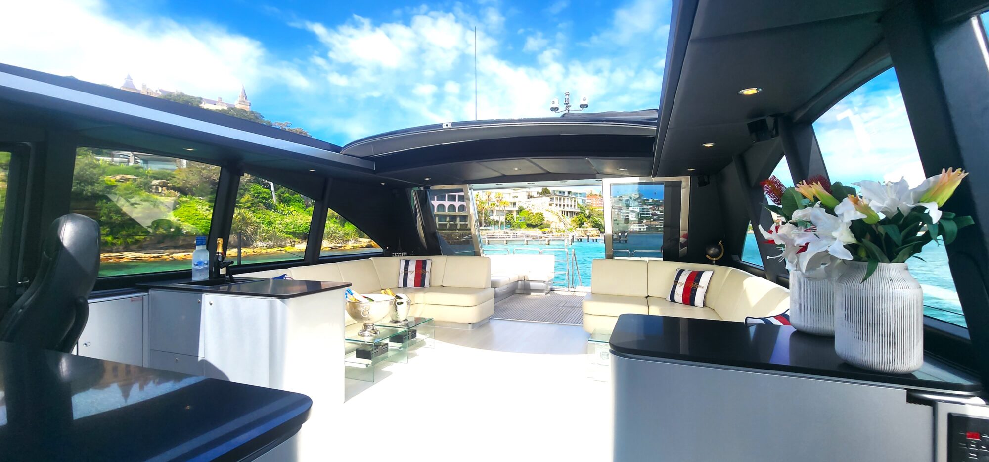 Luxurious yacht interior featuring cream-colored sofas with striped cushions, a bar area, and a vase of lilies. Large windows offer a view of a sunny, coastal backdrop with clear blue skies and lush greenery.