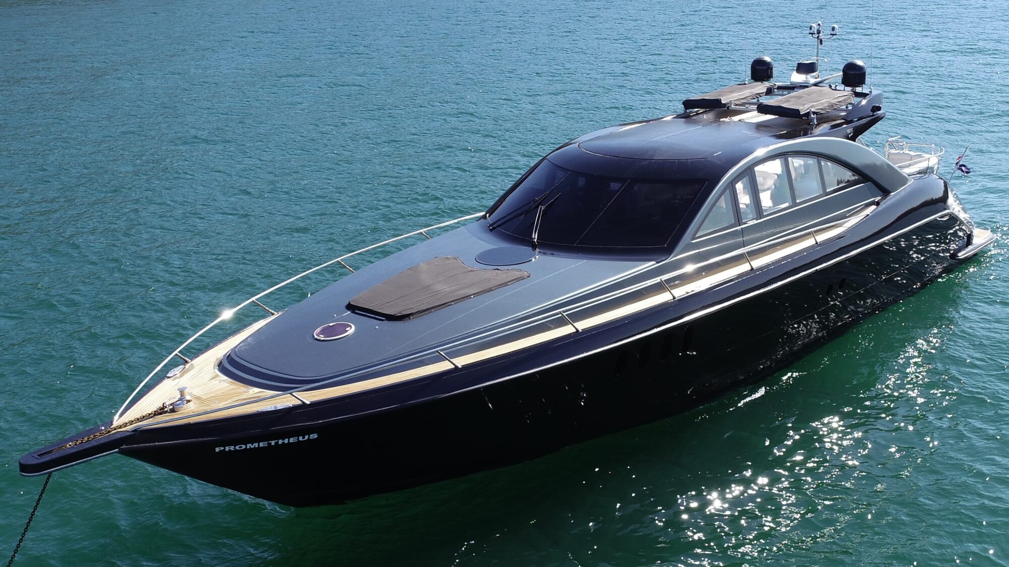 A sleek, modern yacht named "Prometheus" is floating on calm, clear blue water. The yacht features a streamlined design with a black hull and a wooden deck. The sky is clear, suggesting a sunny day.